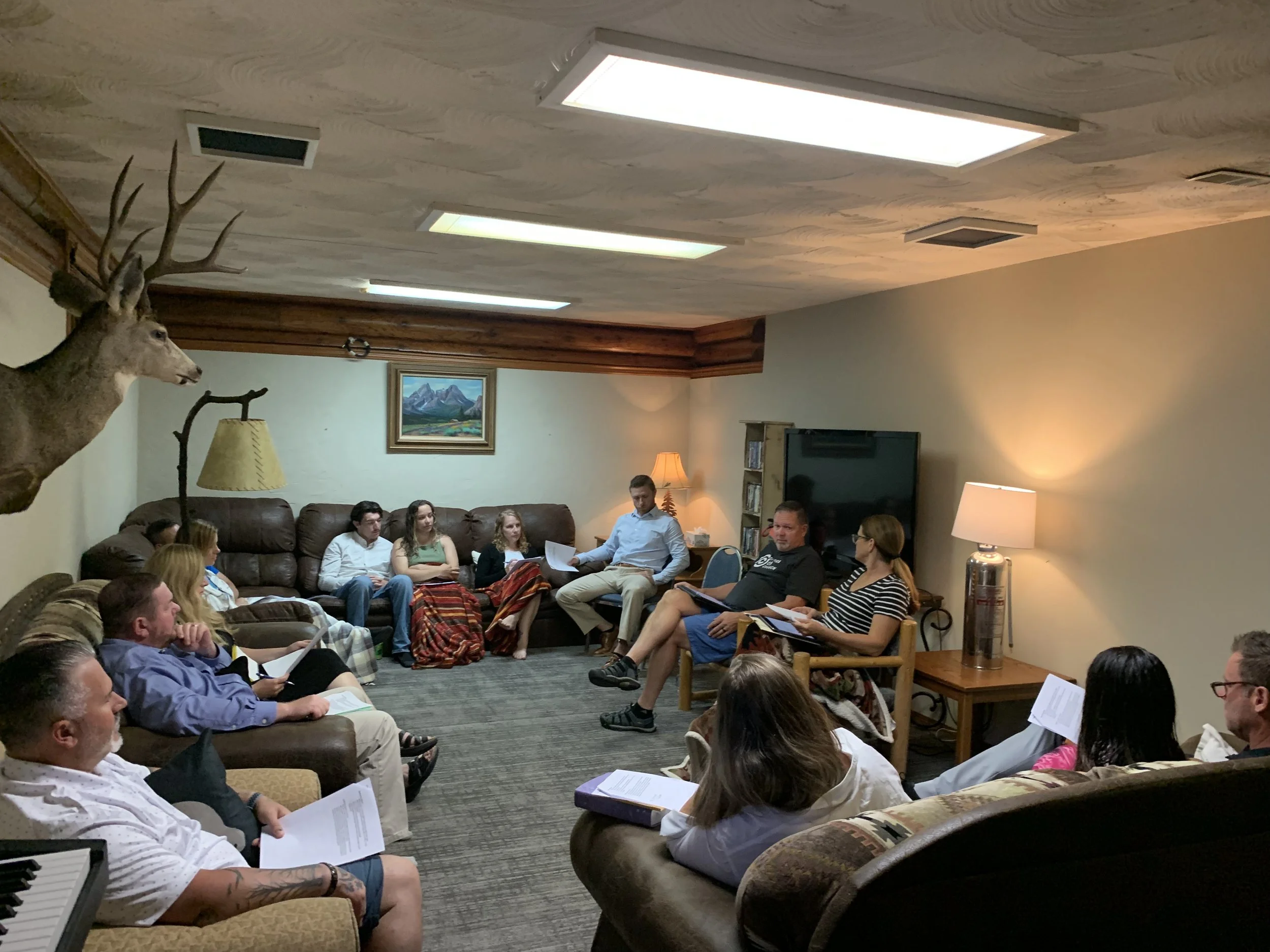 A group of people sitting on couches and chairs in a living room, attending a meeting or discussion, with some holding papers. The room has wood-paneled ceiling, a mounted deer head, a framed landscape picture, and a television set.