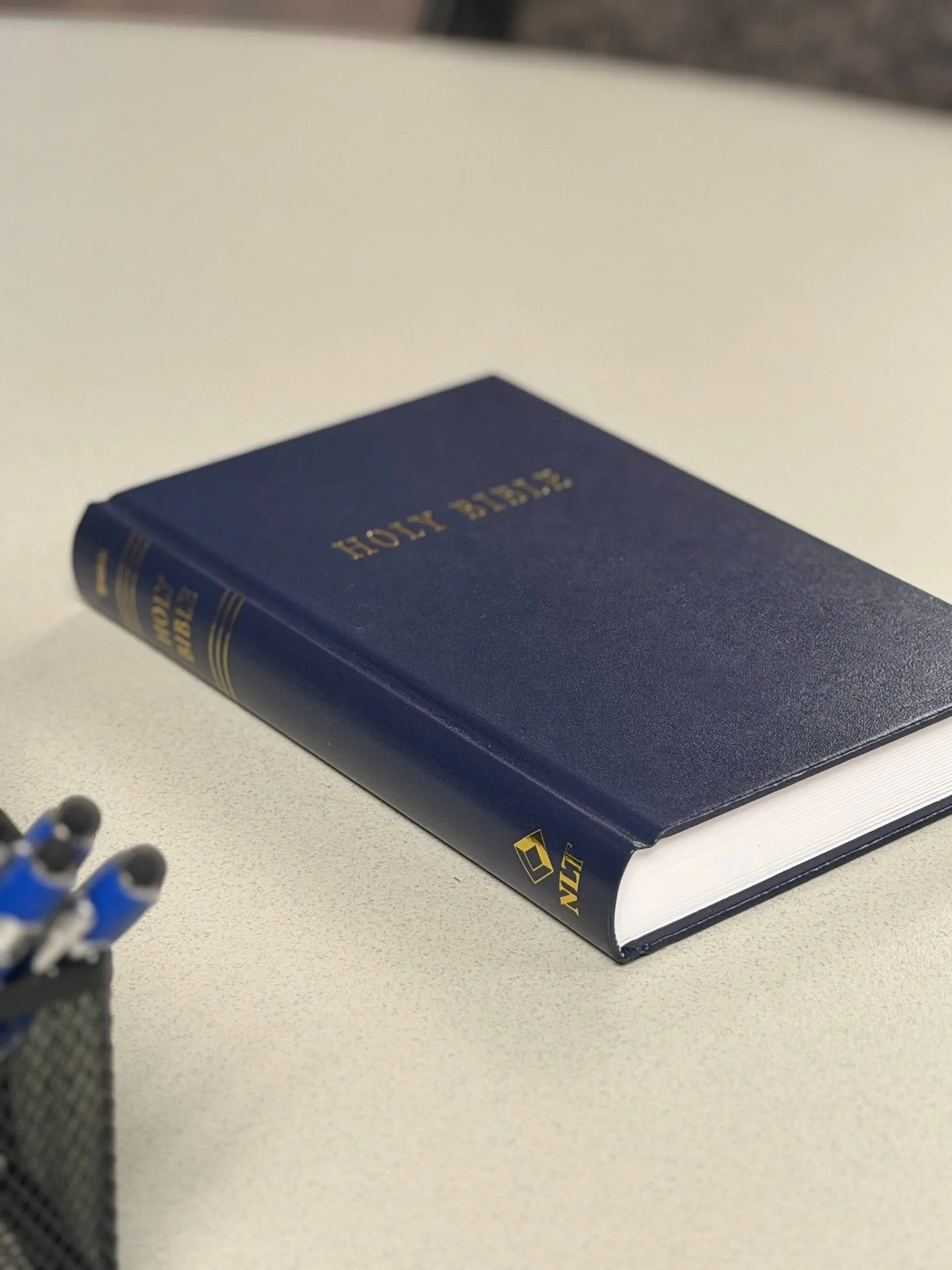 A blue hardcover book titled 'Holy Bible' resting on a light-colored desk or table, with part of a container holding pens or pencils visible in the foreground.