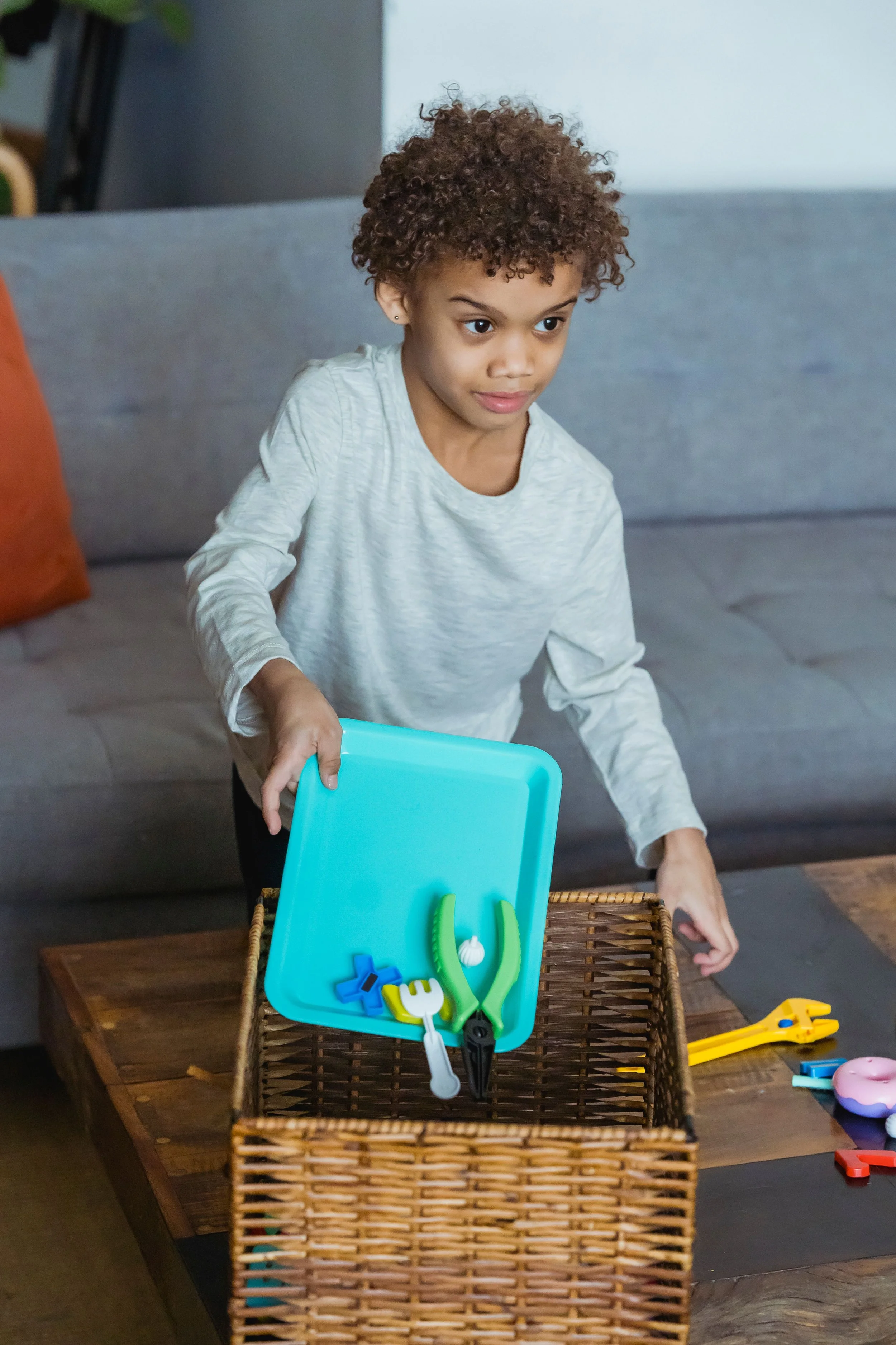 A young girl with curly hair and light skin standing at a table, holding a blue tray with colorful toy utensils, with a wicker basket and children's toys on the table.