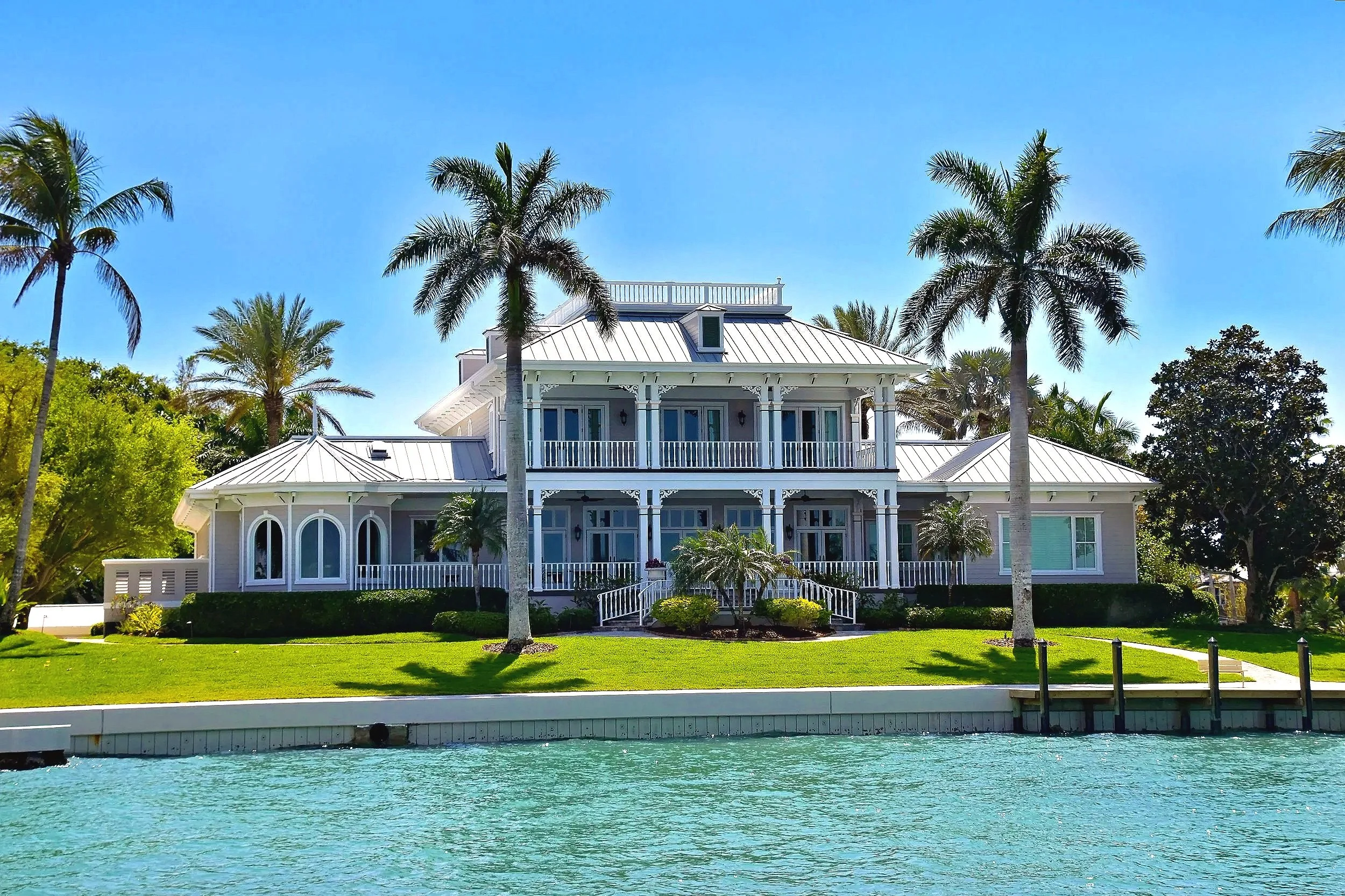 A large, white house with a metal roof, surrounded by palm trees and greenery, situated along a body of water under a clear blue sky.