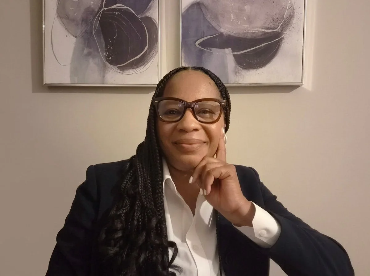 A woman with braided hair and glasses, dressed in a white shirt and dark blazer, sitting in front of abstract artwork with dark and light circles on a wall.