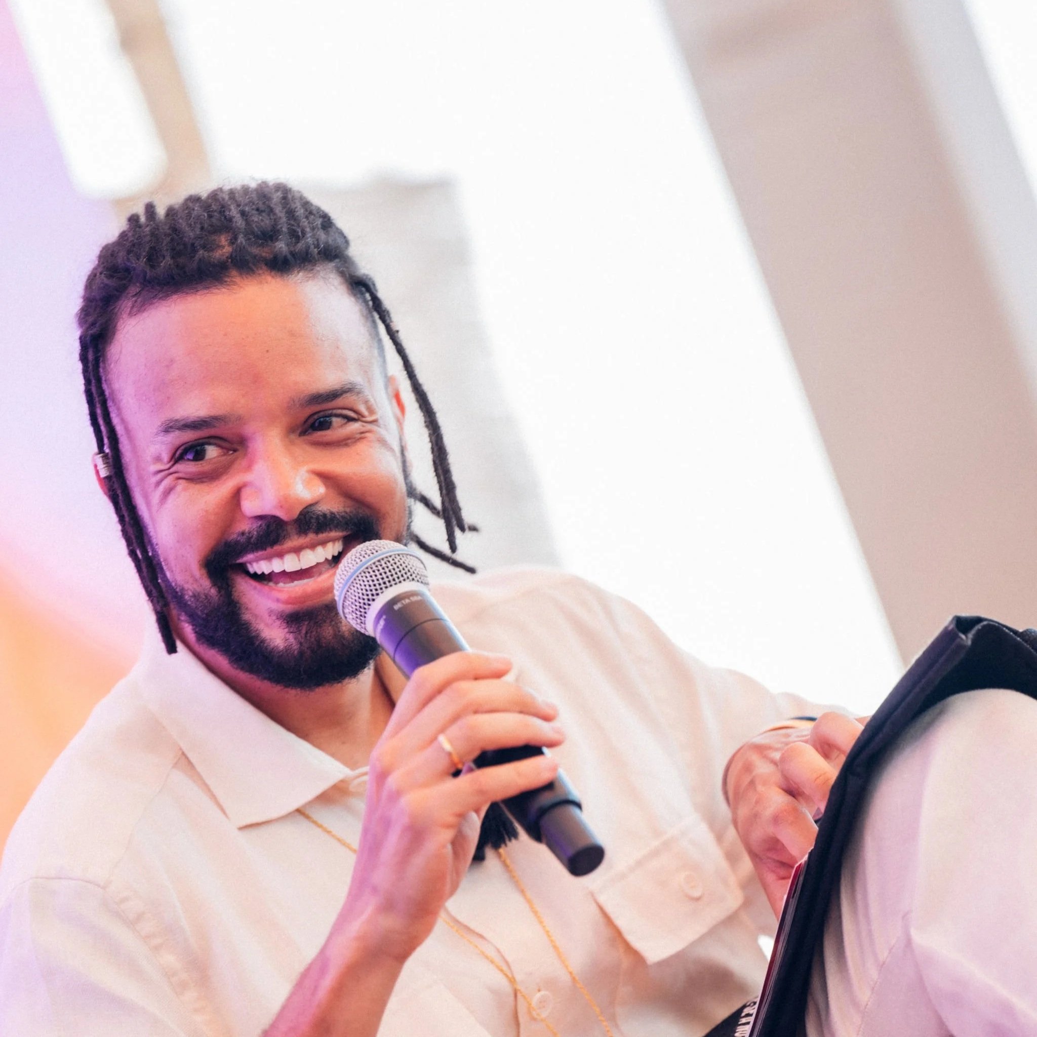 A person with dreadlocks smiling and holding a microphone. The individual is wearing a light-colored shirt and appears to be speaking or singing. The background is plain and softly lit.