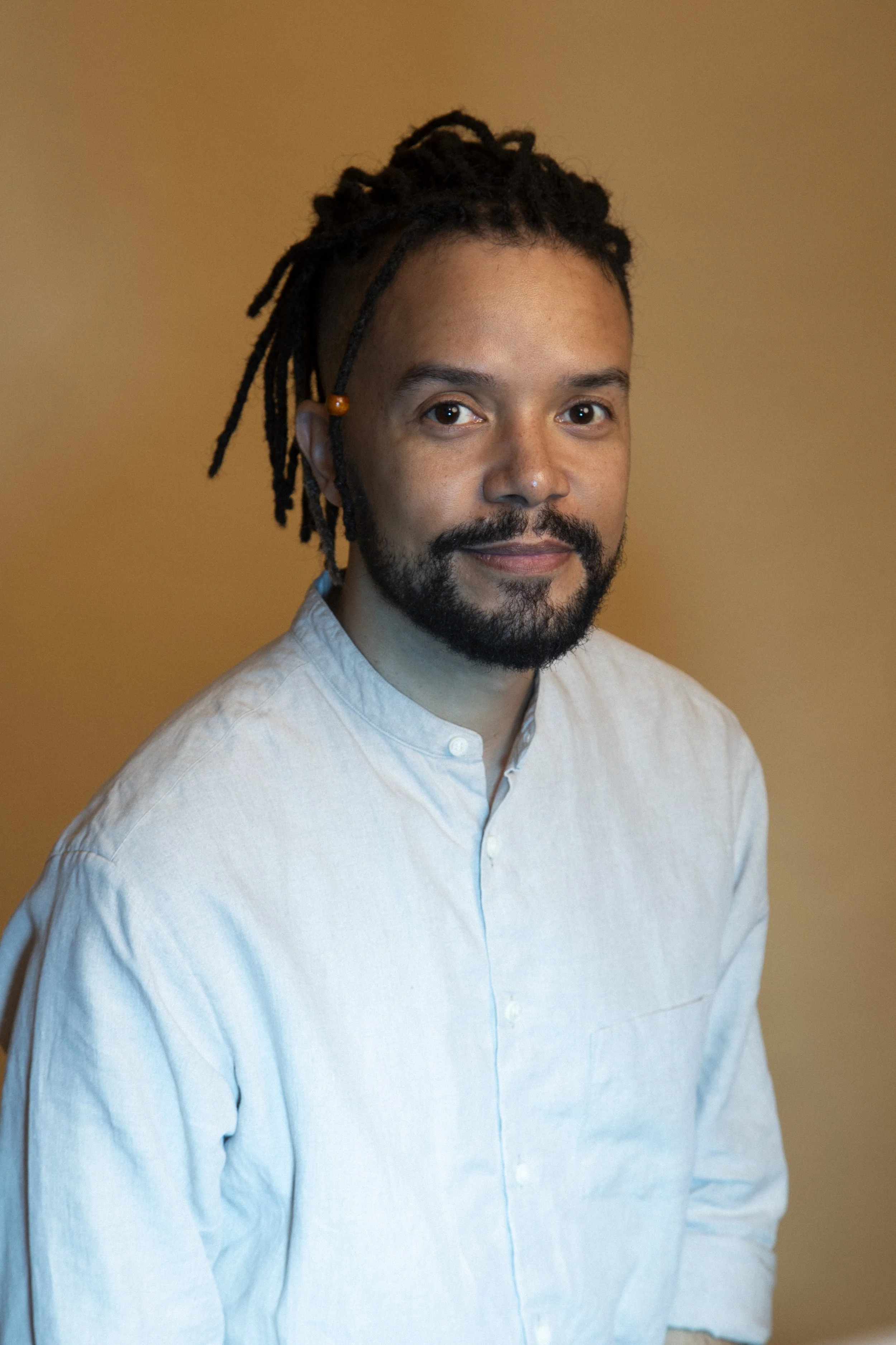 A man with short hair, a beard, and dreadlocks with beads, wearing a white button-up shirt, looking at the camera with a neutral expression, standing against a plain brown background.