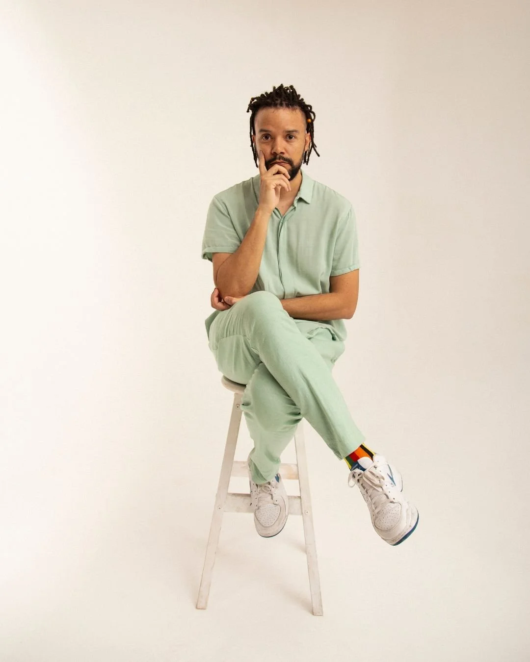 Man with dreadlocks sitting on a white stool against a plain white background, wearing a light green shirt and pants, white sneakers, and colorful socks, with his hand on his chin in a thoughtful pose.