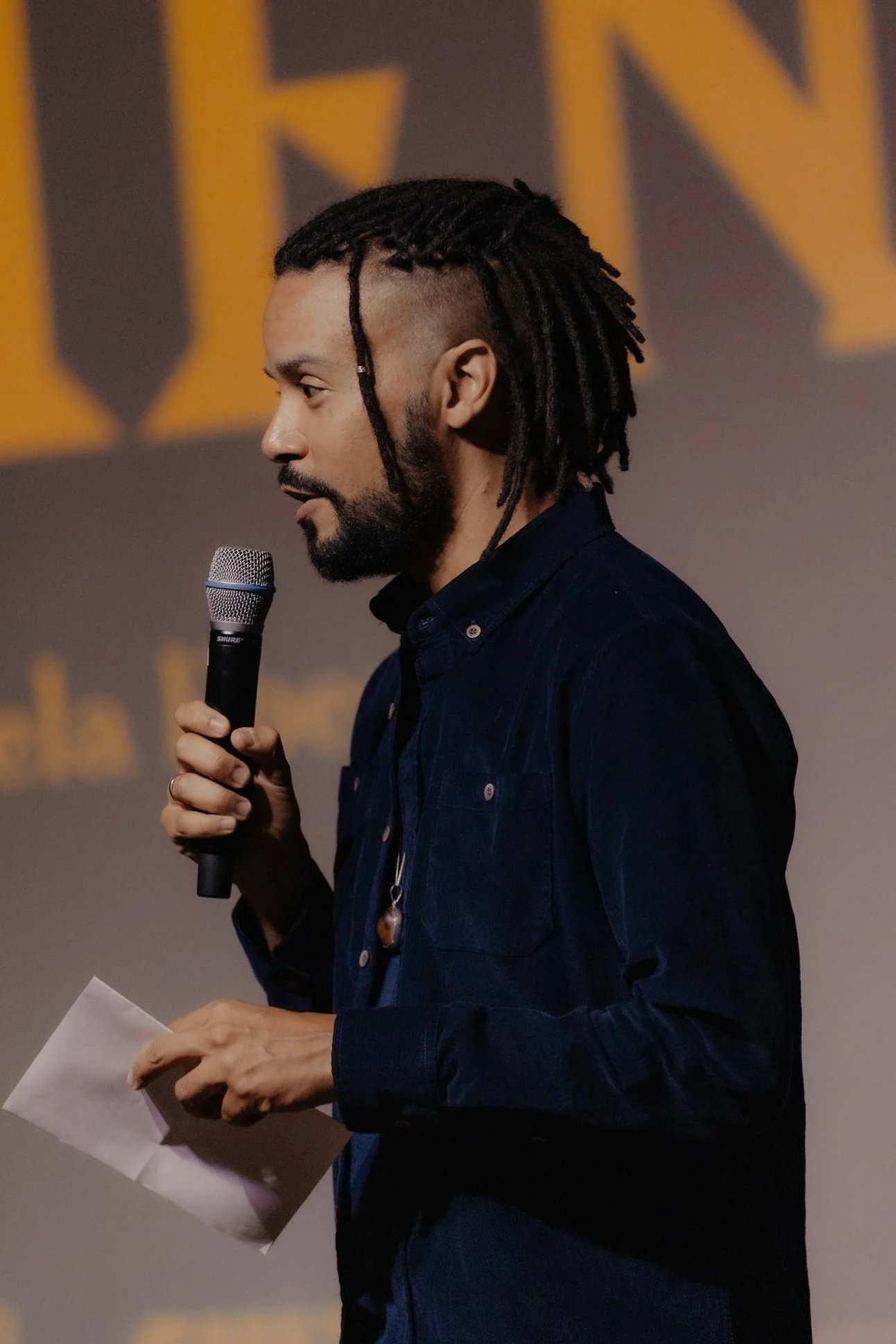 A man with long dreadlocks and a beard is speaking into a microphone. He is wearing a dark jacket and holding some papers.