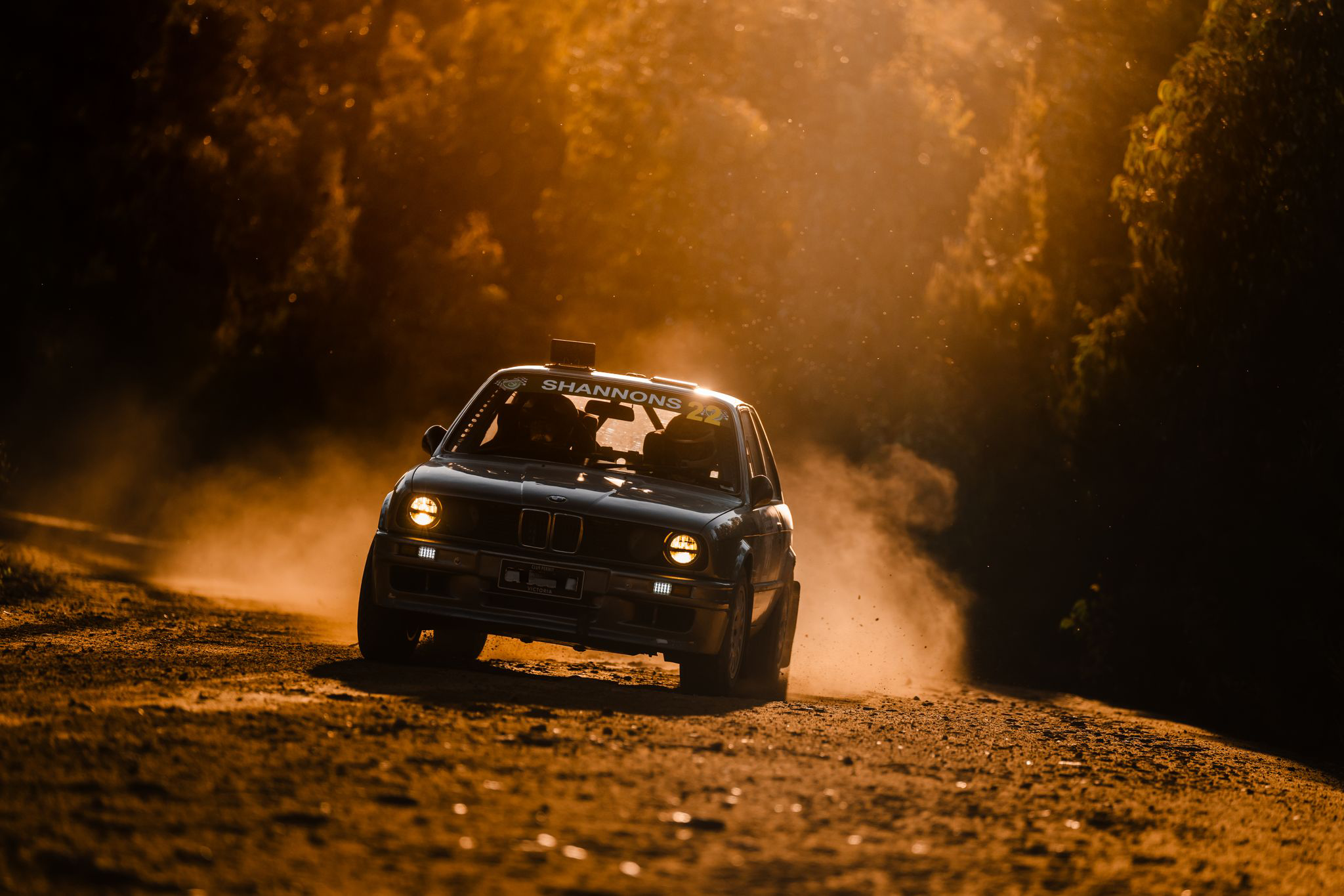 A black BMW race car with yellow headlights is driving on a dirt track at sunset, kicking up dust behind it.