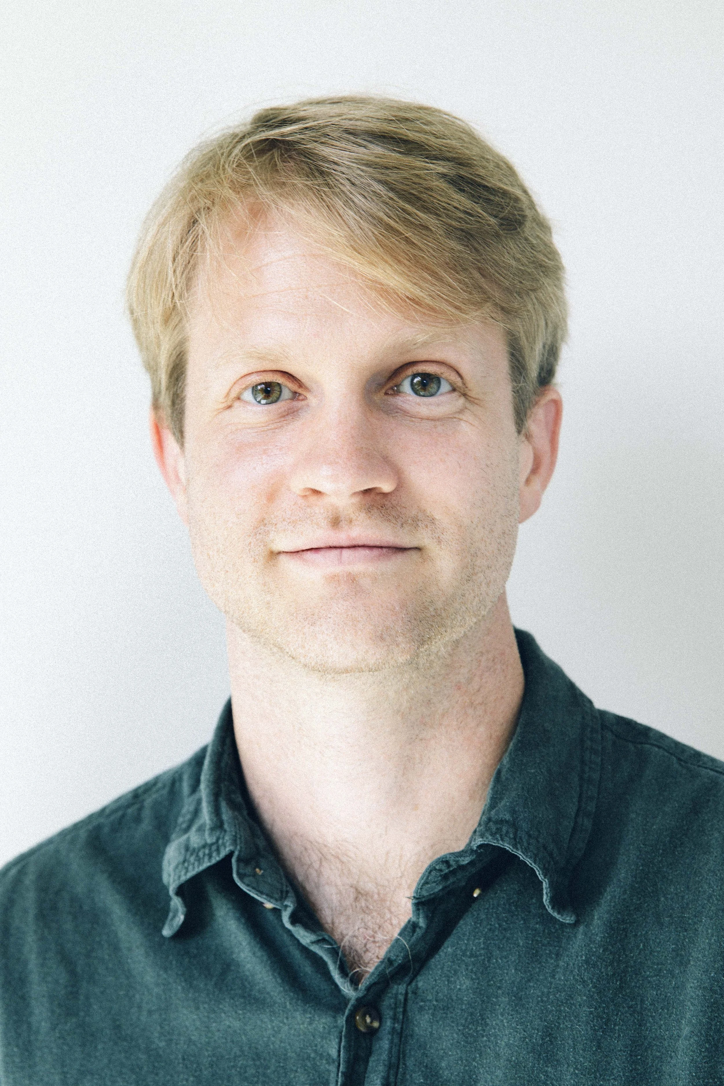 Close-up portrait of a young man with light skin, short blond hair, green eyes, wearing a dark green button-up shirt, against a light background.