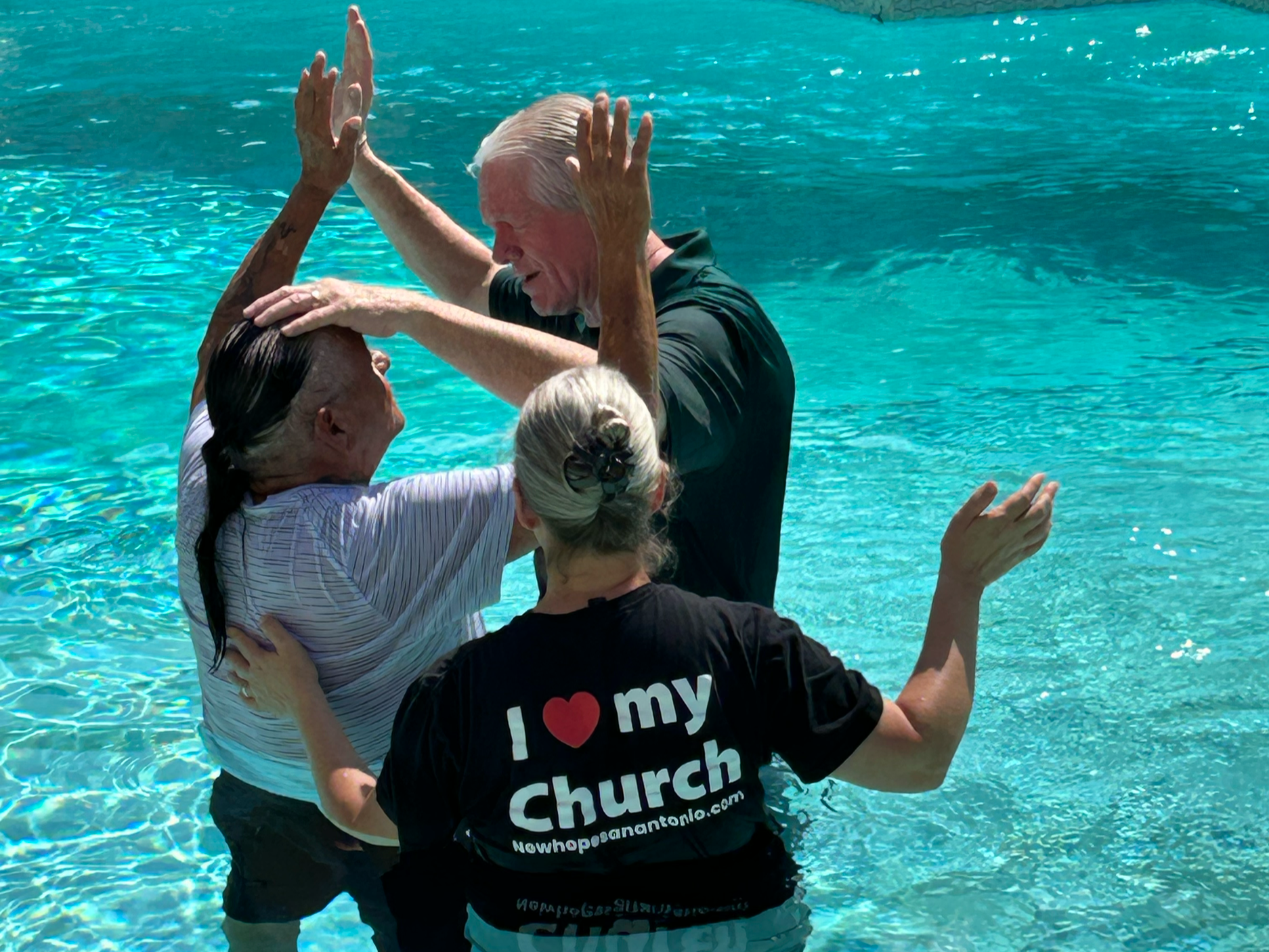 Pastor and Pastors' Wife stand in a pool, all three of them with hands raised, praying over a man who was just baptized. 