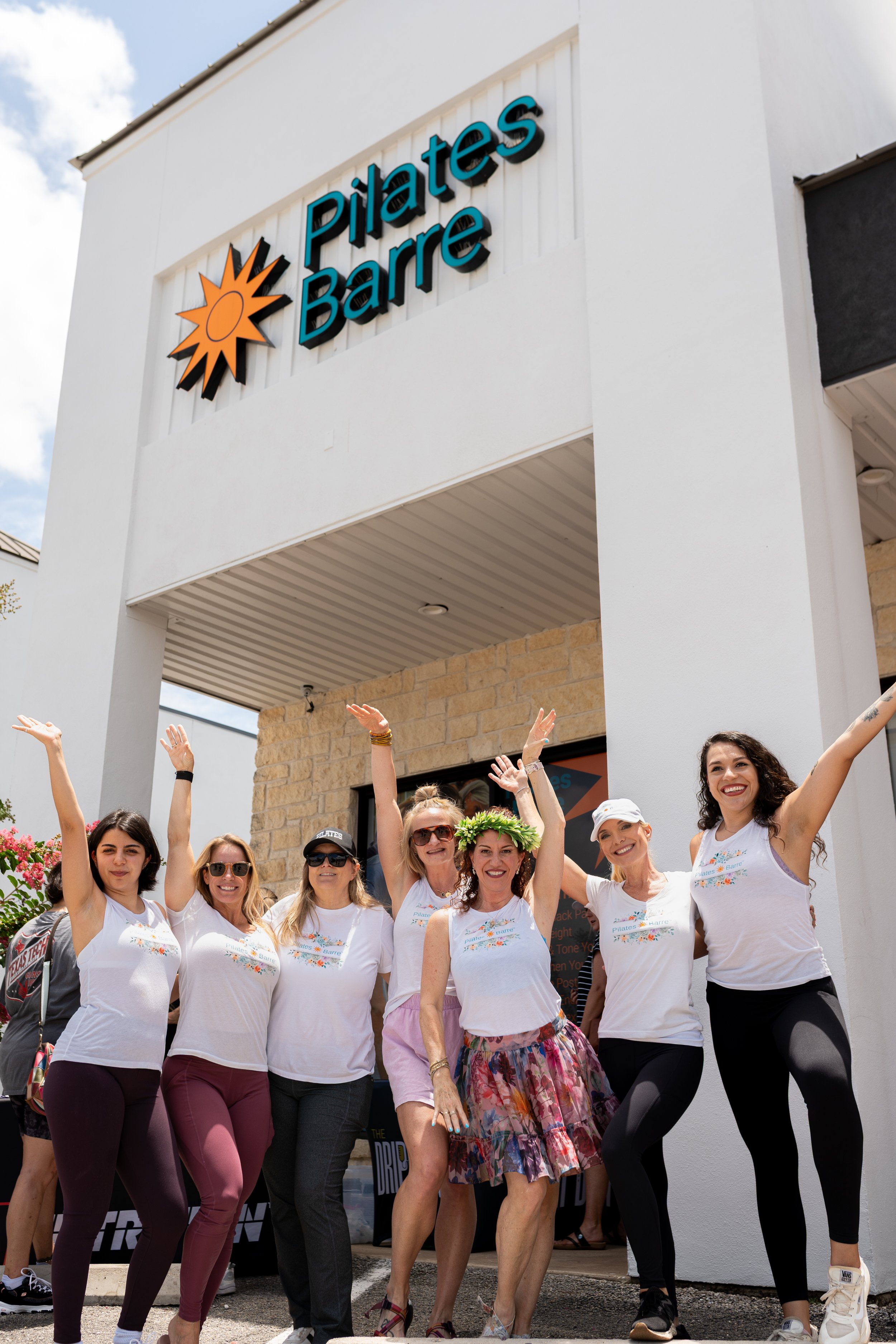 A group of women celebrating outside a Pilates studio called Pilates Barre, with arms raised and smiling, some wearing matching white T-shirts with a floral logo.