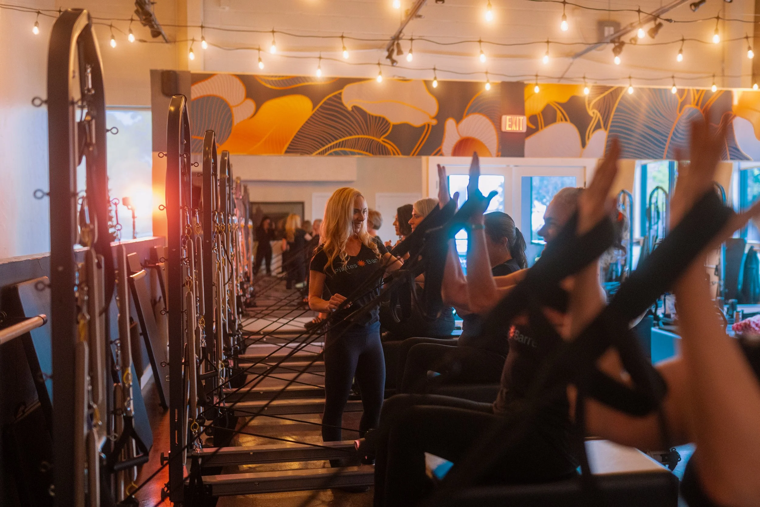 A group of people participating in a Pilates class inside a warmly lit studio with string lights and a large decorative mural on the wall. The participants are seated and using resistance bands for exercise, with an instructor guiding them.