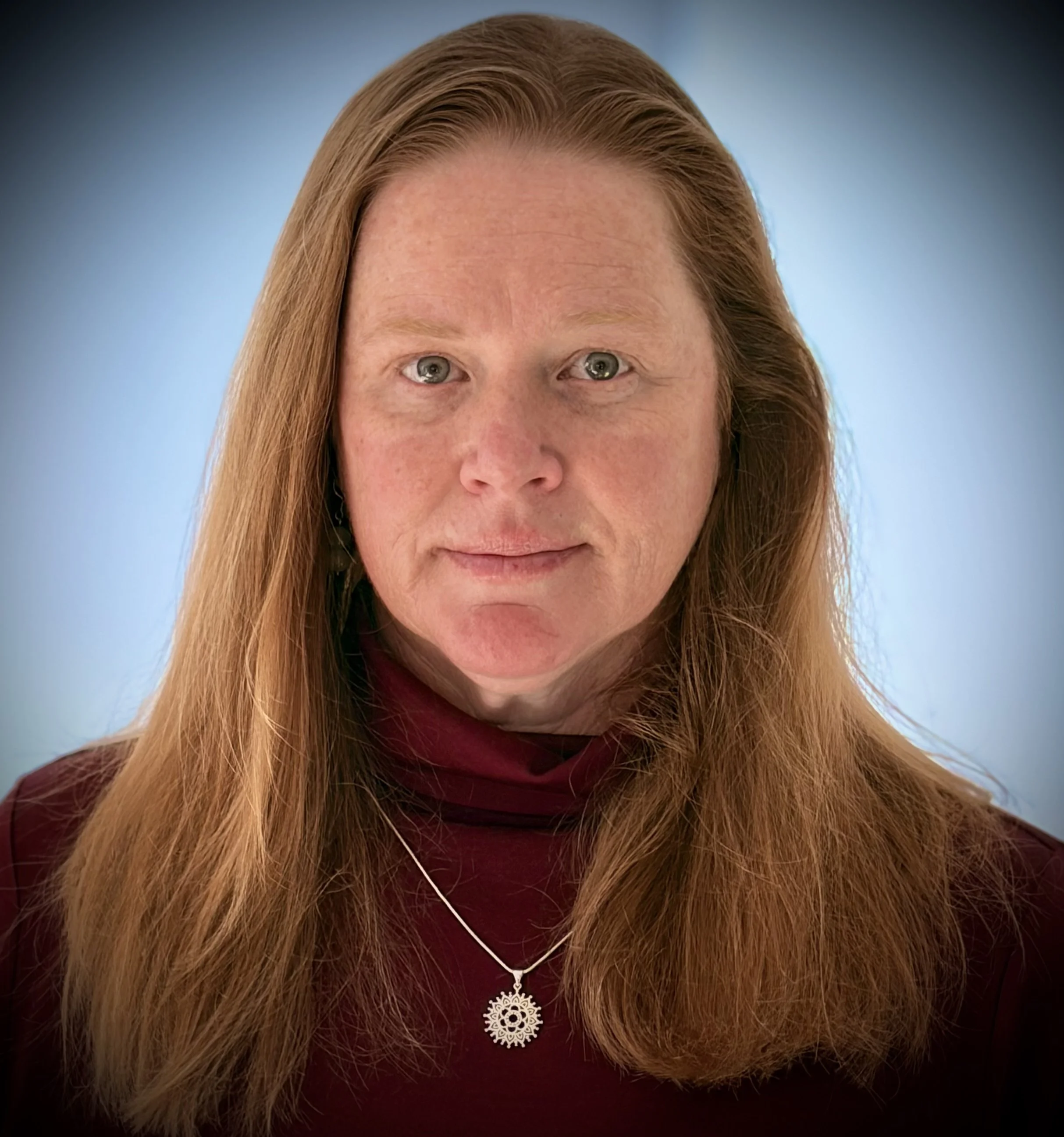 Close-up of a woman with long red hair wearing a black top and a silver pendant necklace, against a soft blue background.