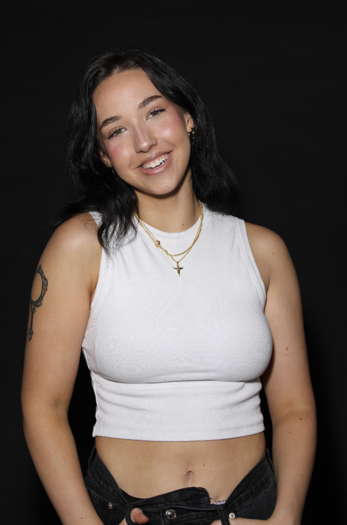 A young woman with black hair, wearing a white sleeveless crop top, black jeans, and jewelry, smiling against a black background.