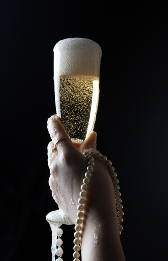 A hand holding a champagne flute with bubbles and foam at the top, against a dark background, with a pearl necklace draped around the hand.