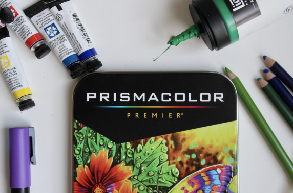 A Prismacolor Premier colored pencils tin box surrounded by art supplies including paint tubes, a green paint bottle, and colored pencils on a white surface.