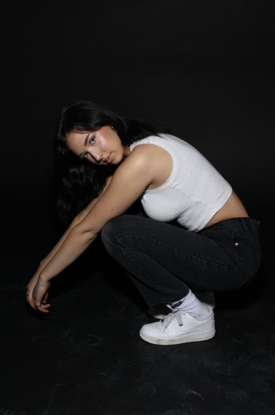 A young woman with long dark hair squats against a black background, wearing a white tank top, black jeans, white socks, and white sneakers, looking at the camera.