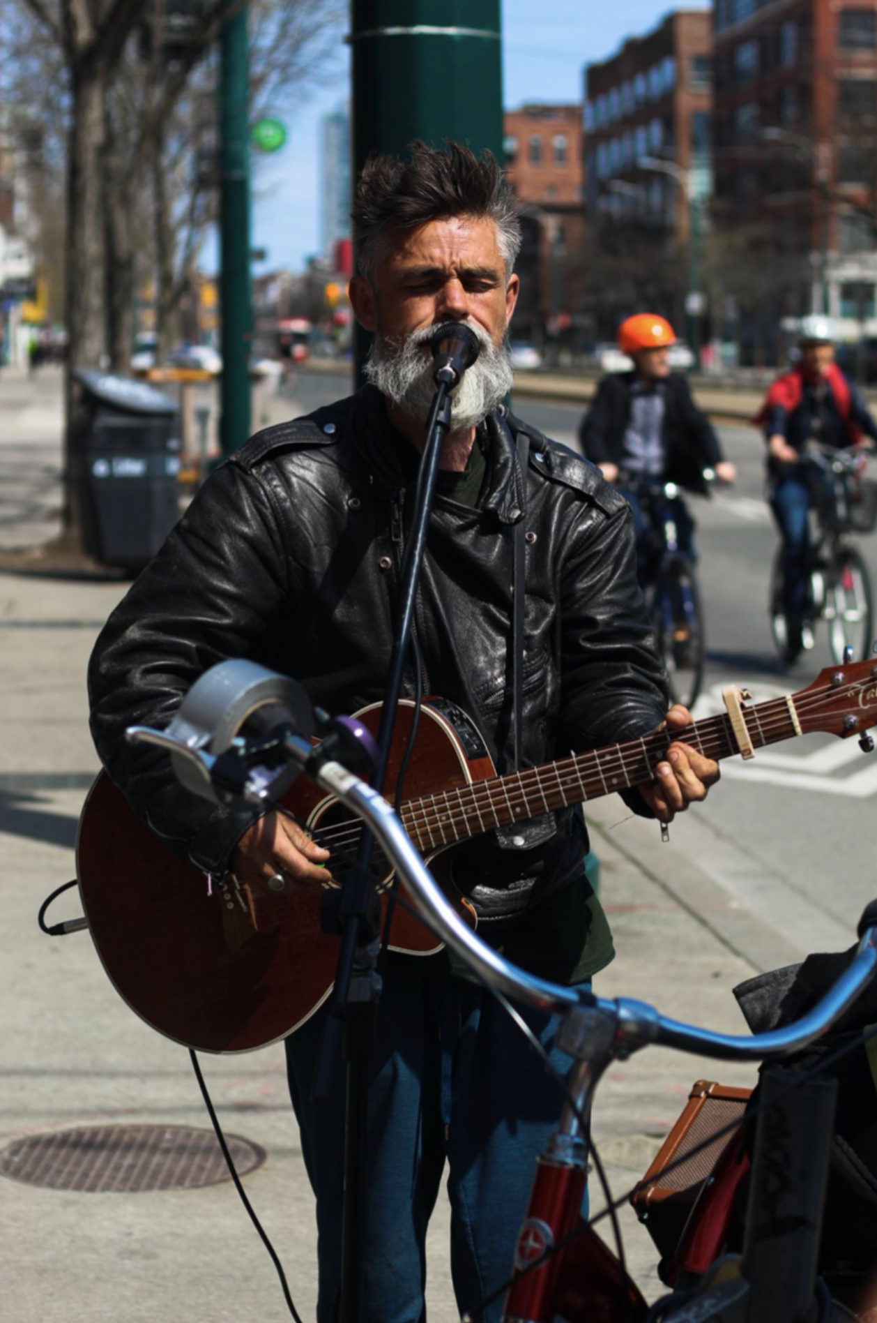 A man with a gray beard and dark hair plays an acoustic guitar and sings into a microphone on a city sidewalk, with cyclists and brick buildings in the background.