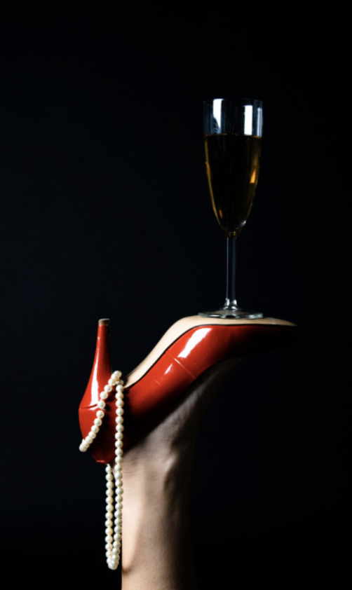 A woman's hand with a red high heel shoe on the finger, strands of pearl necklace draped over the shoe, a champagne flute filled with champagne on the shoe's heel, against a black background.
