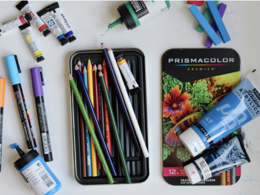 Art supplies including colored pencils, markers, watercolor tubes, and crayon box on a white surface.