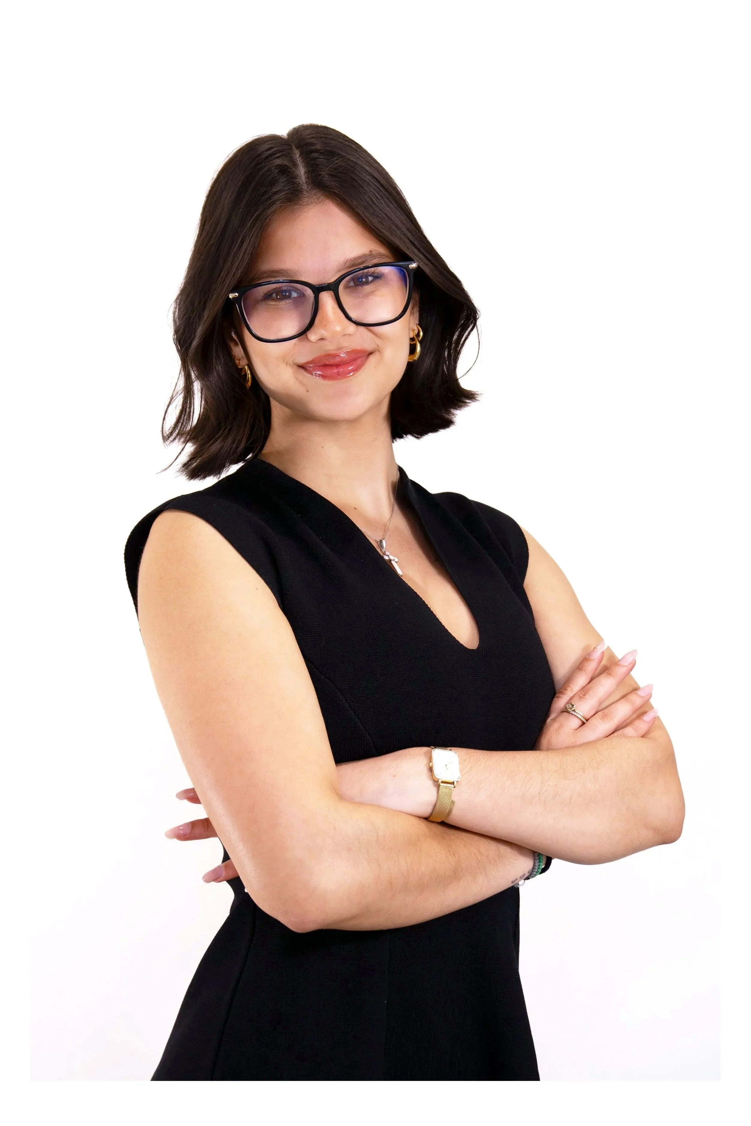 A woman with shoulder-length dark hair, wearing large black-rimmed glasses, a black sleeveless dress, gold hoop earrings, a gold wristwatch, a silver necklace, and a ring on her right hand, smiling and crossing her arms against a white background.
