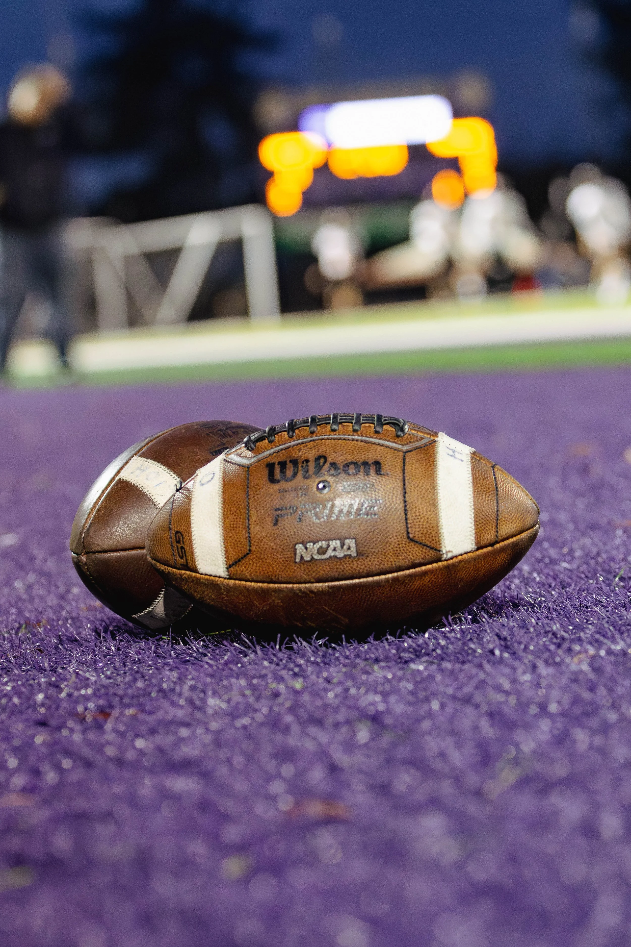 A football resting on a purple field with blurred stadium lights in the background.