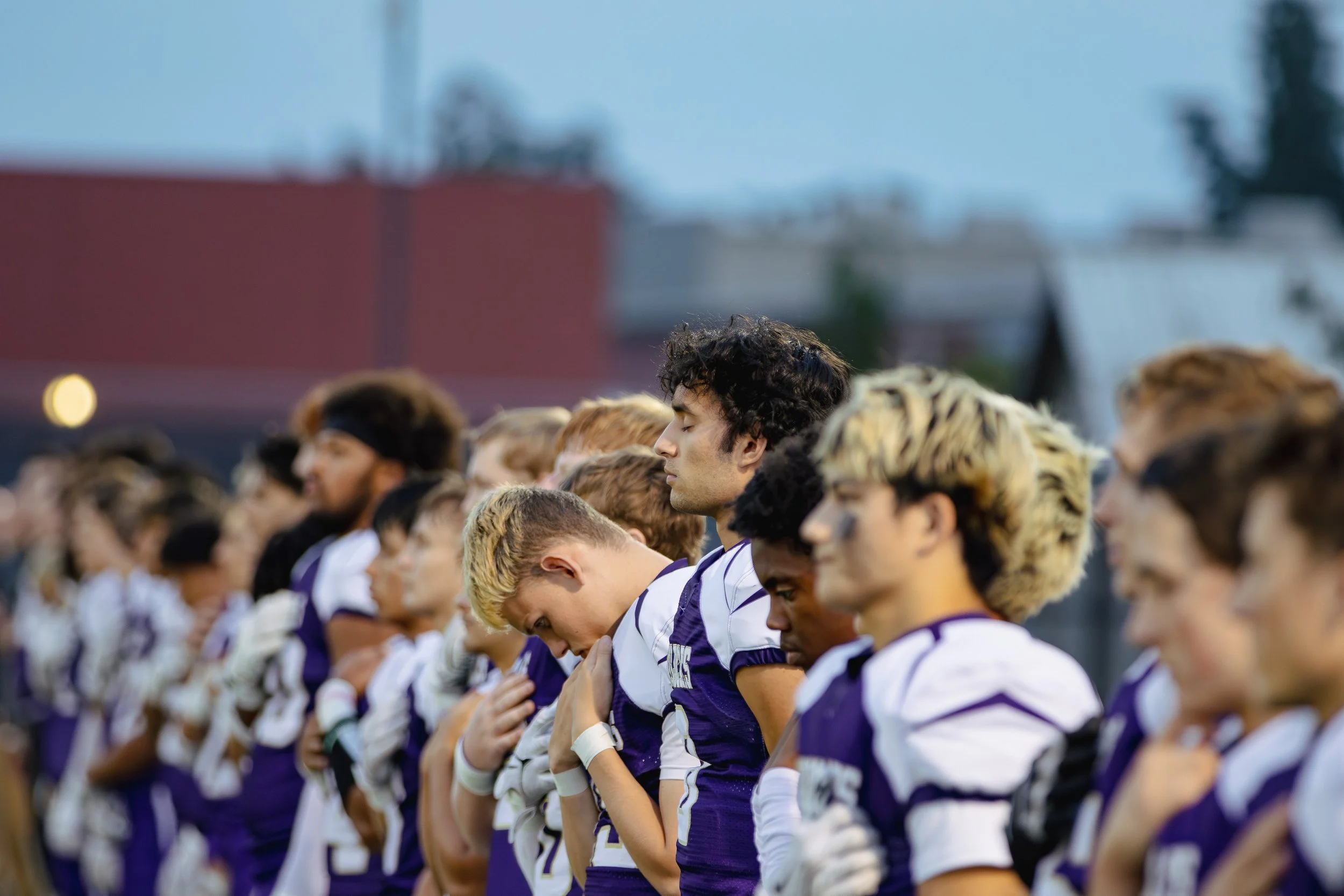 A group of high school students standing with their hands over their hearts during a patriotic moment, wearing purple and white sports jerseys, with the sky and some rooftops in the background.
