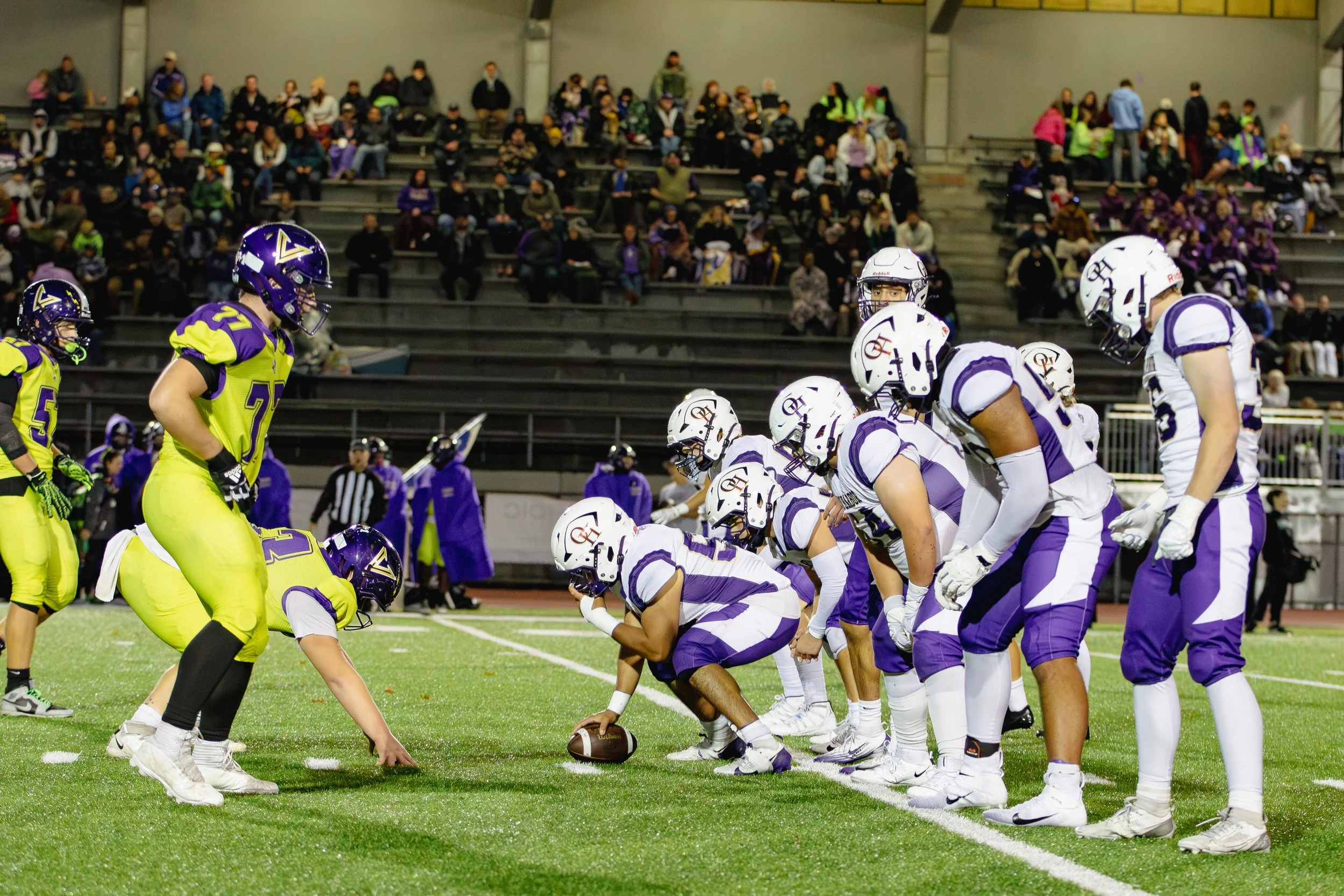 A football game is in progress with players in white and purple uniforms facing off against players in bright yellow uniforms. The players are on a green field, with spectators sitting in the bleachers in the background.