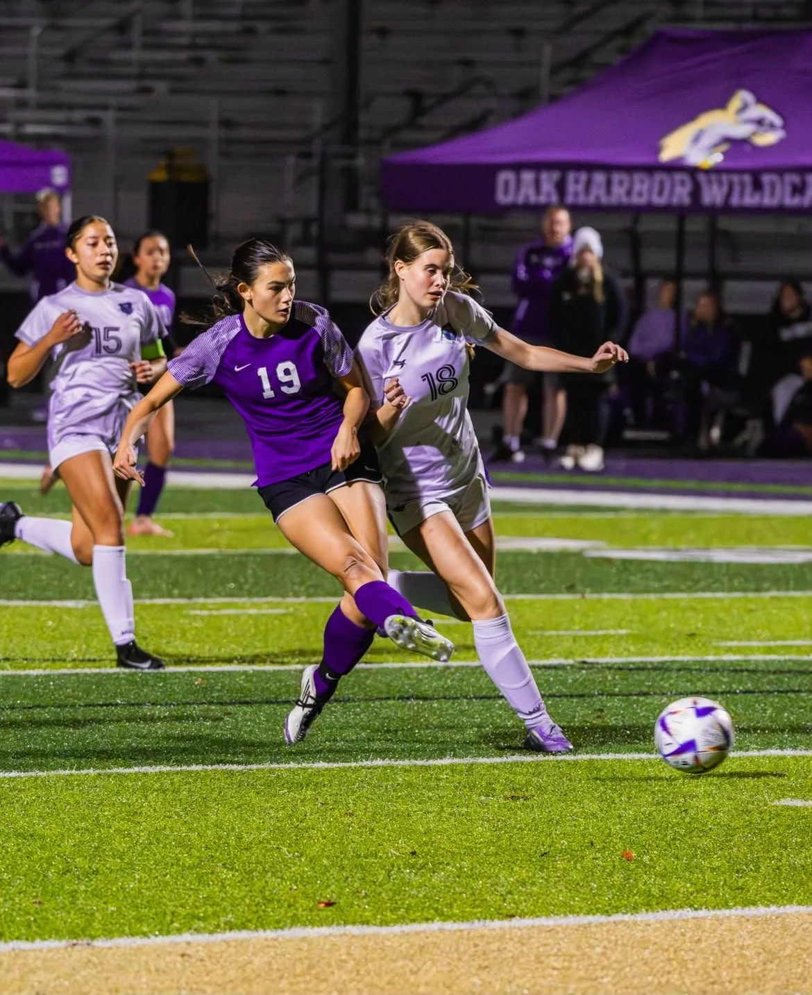 Two female soccer players are chasing the ball on a field during a game at night. One player in a purple jersey with number 19 and purple socks is kicking the ball, while another player in a white jersey with number 18 and white socks is running alongside her. Other players and spectators are visible in the background.