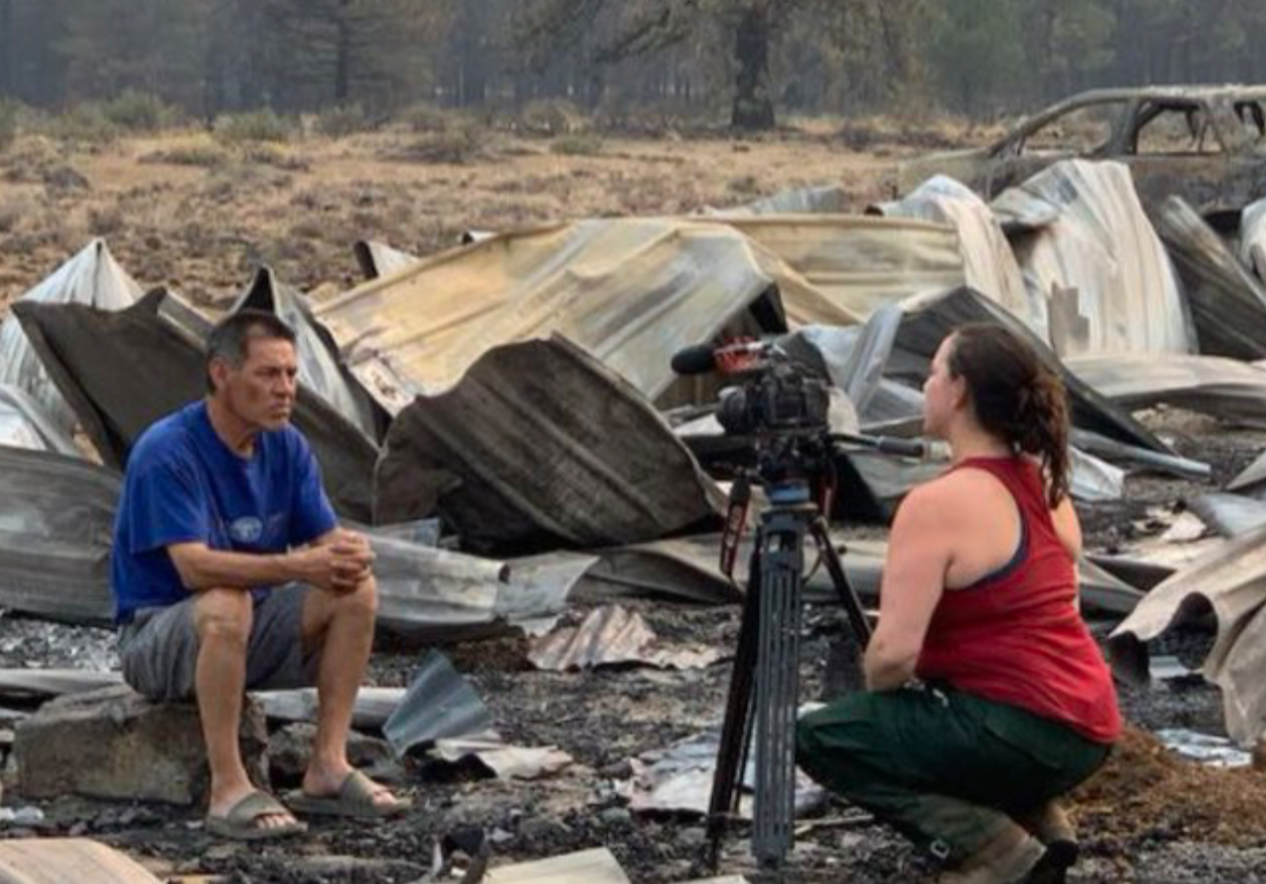 A woman in a red tank top and camo pants being interviewed by a man in a blue shirt while sitting amidst the charred remains of a destroyed area, likely after a wildfire.