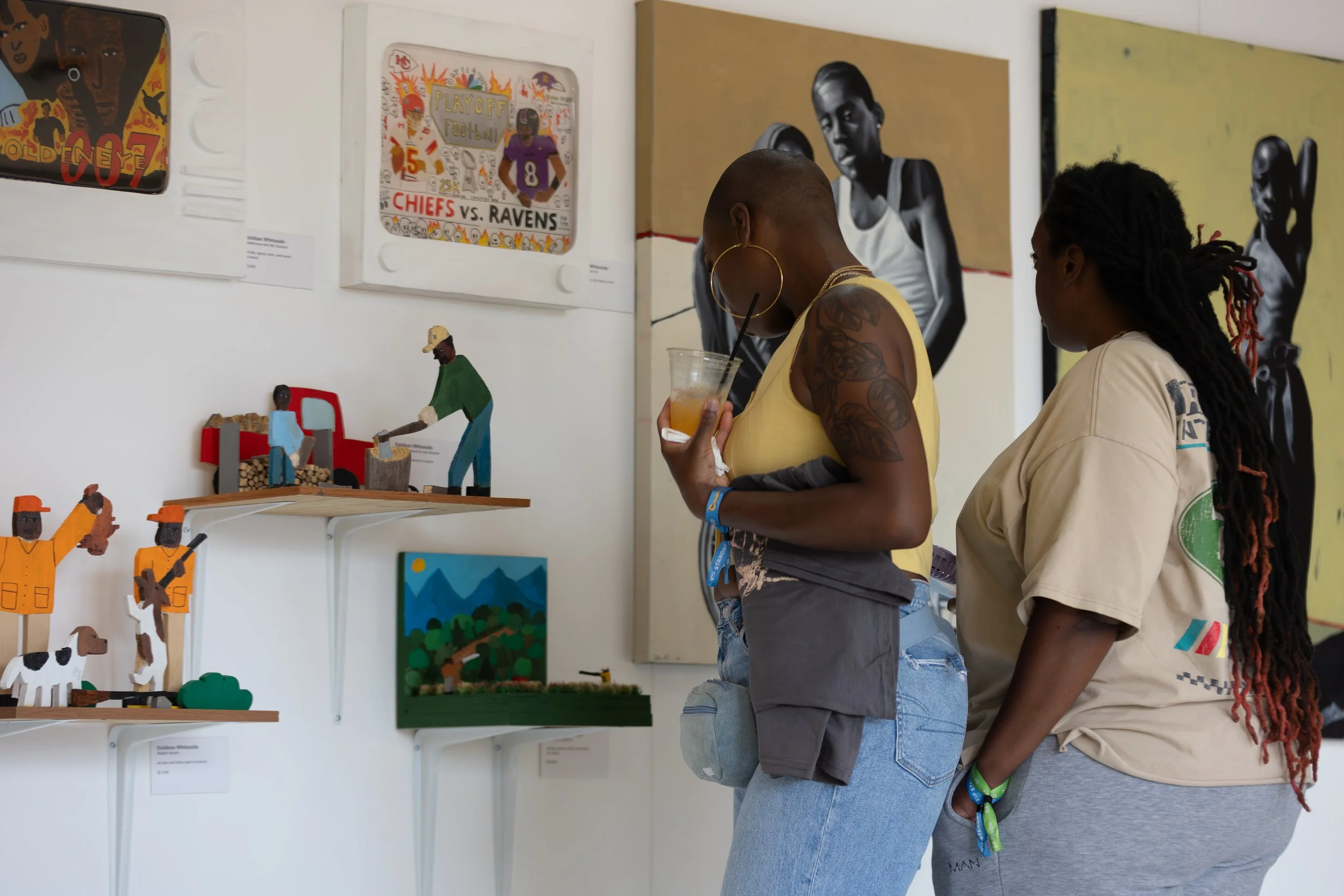Two women are looking at artwork displayed on a wall in an art gallery. The art includes colorful paintings and handcrafted wooden figures. One woman is wearing a yellow top and has large hoop earrings, holding a drink, and the other woman has braided hair and is wearing a light-colored jacket.