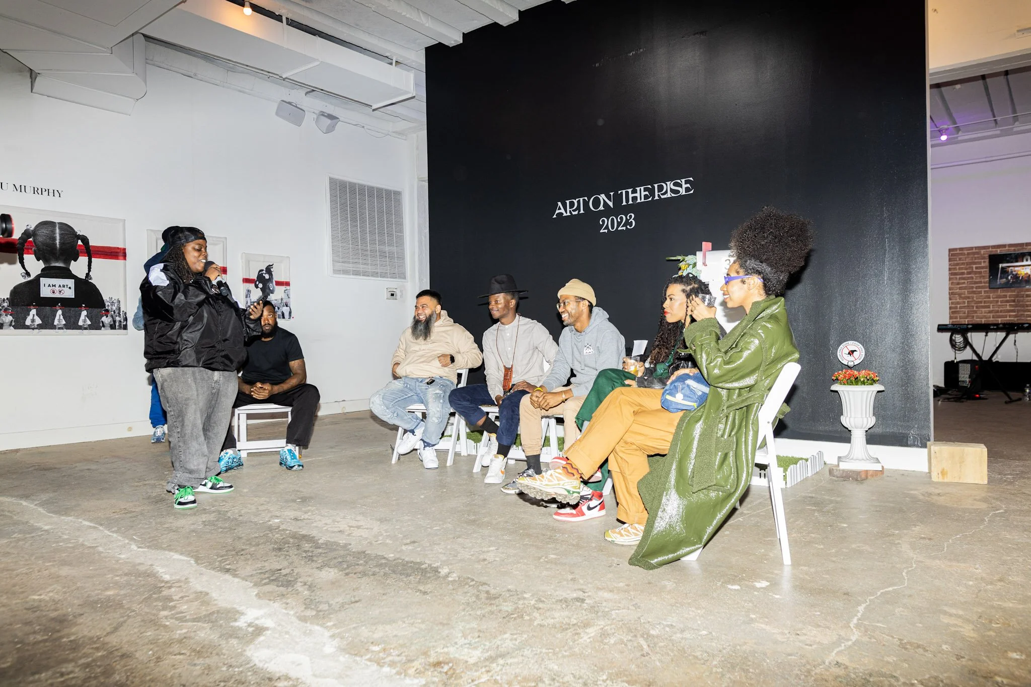 Group of nine people sitting in chairs on a stage, with a woman standing and speaking into a microphone. The event appears to be an art presentation or discussion, with a black wall in the background displaying 'ART ON THE RISE 2023.'