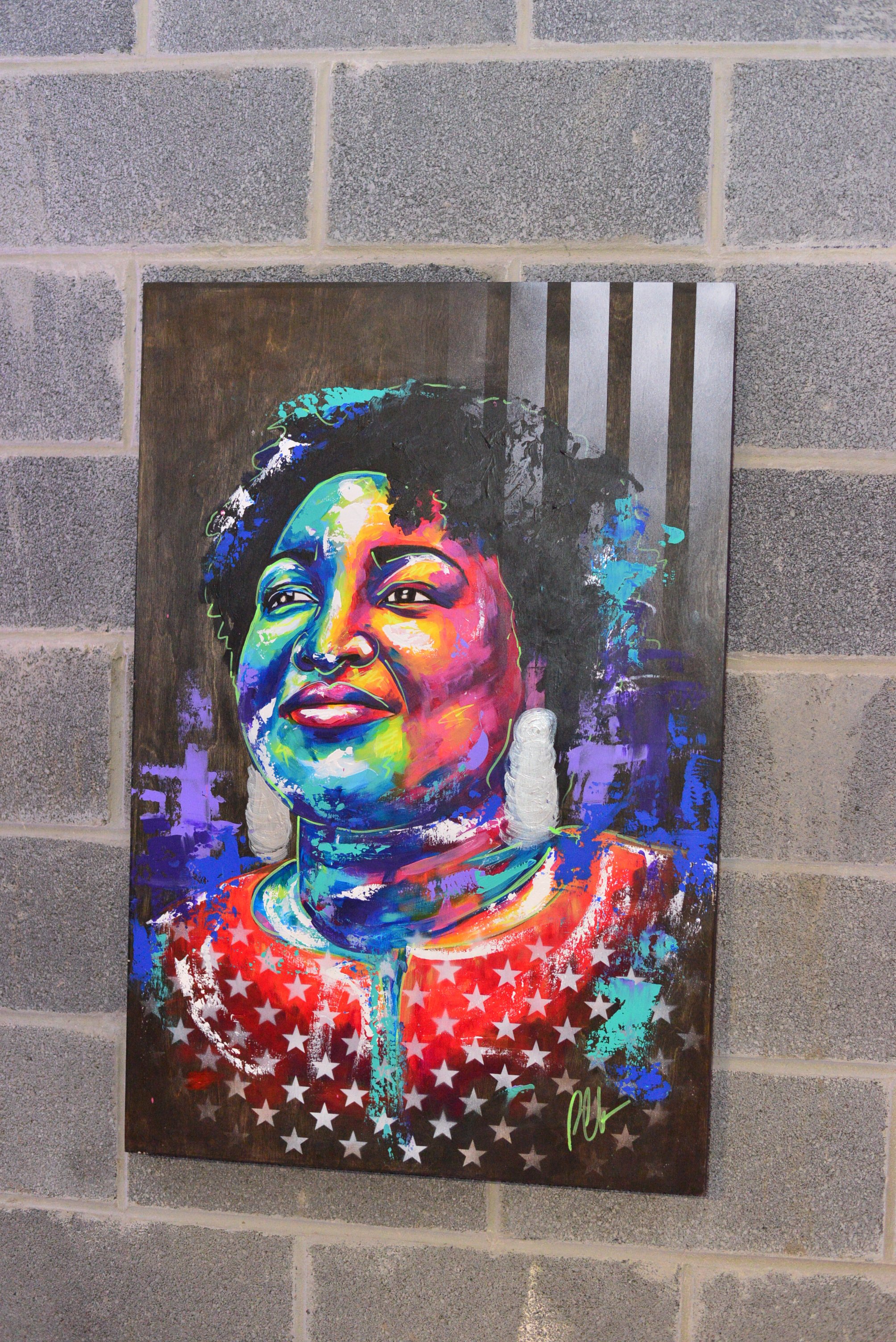 A colorful portrait of a woman with natural hair, painted in vibrant colors on a dark wooden background, with a red, white, and blue star pattern at the bottom and a gray and black striped pattern at the top right.
