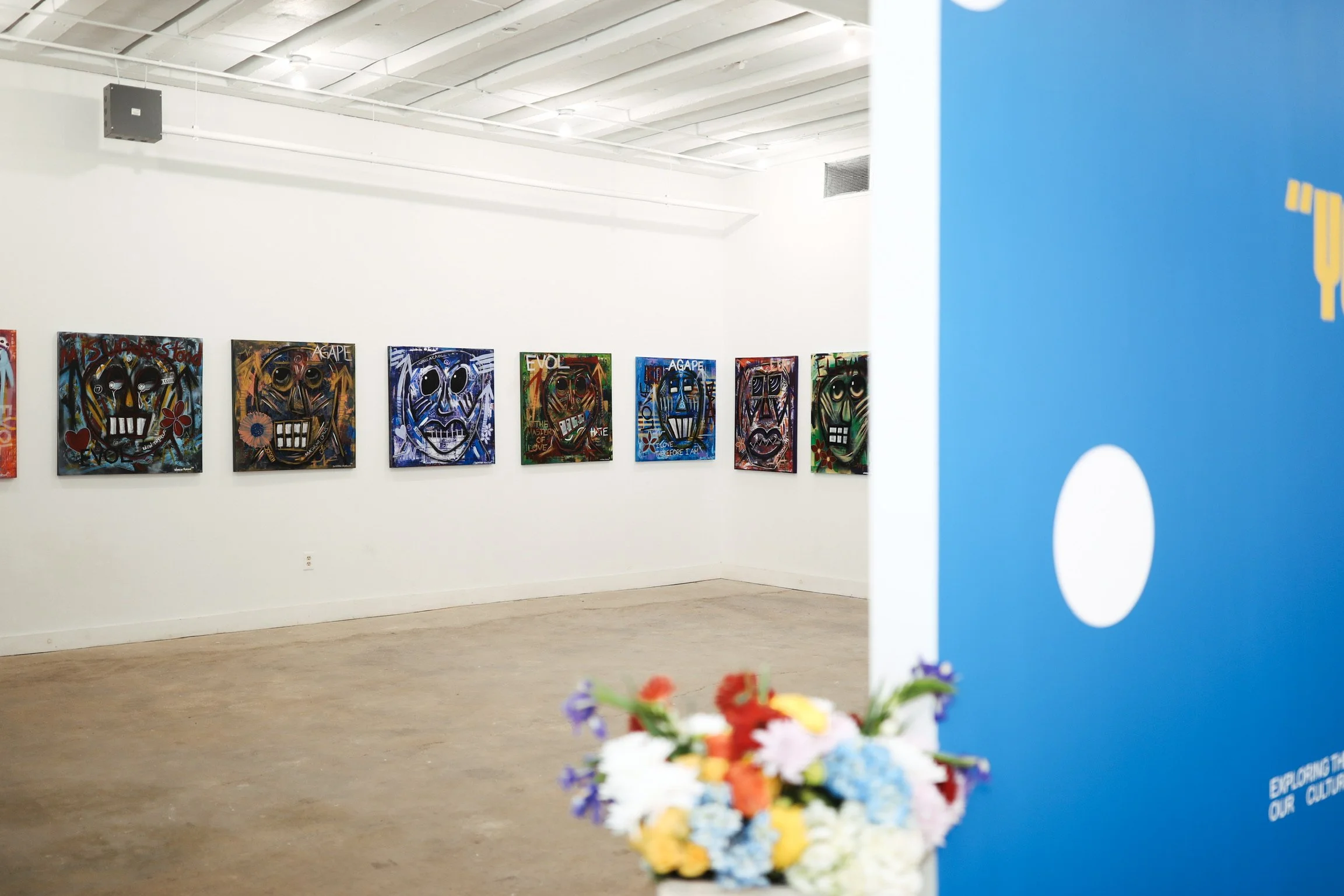 Art gallery with colorful abstract portrait artworks of faces on a white wall, and a blue banner on the right side with yellow and white design elements partially visible, and a blurred flower arrangement in the foreground.