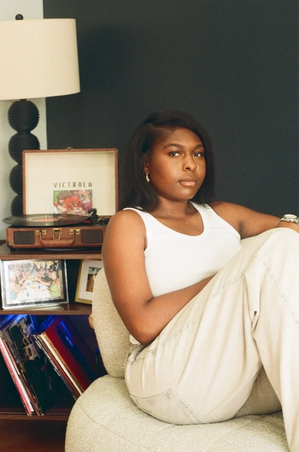A young woman with dark hair and wearing a white tank top and beige pants is sitting on a cream-colored chair in a living room. Behind her is a dark wall with a lamp, a record player, and framed photos, including one of a colorful parade and one of a birthday celebration.