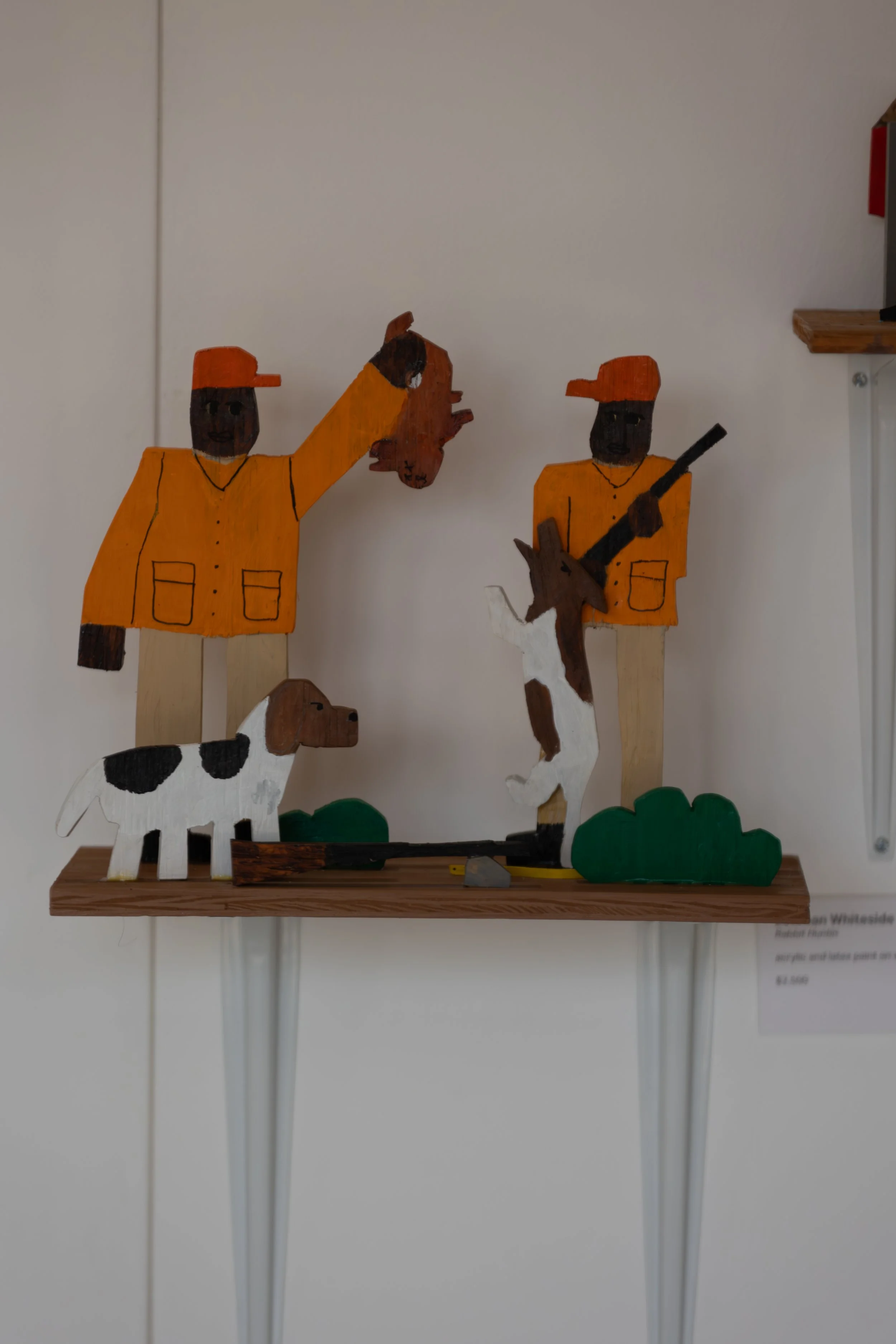 Wooden painted figures of two hunters, a dog, and a deer scene on a wooden shelf.