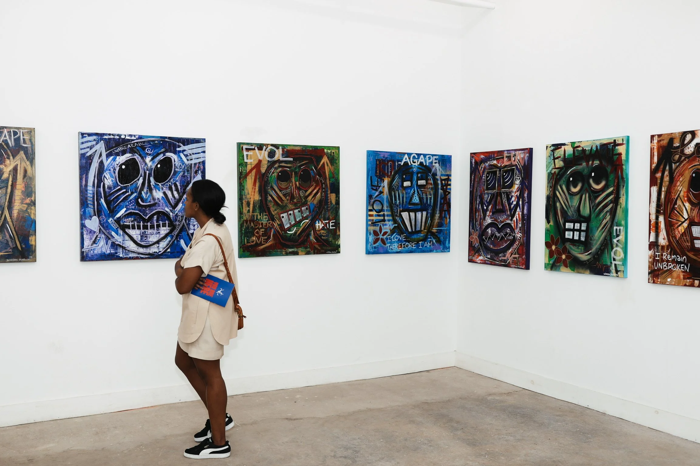 A woman with dark hair tied back, wearing a beige outfit and black sneakers, standing in an art gallery, looking at colorful abstract paintings of skulls on the white wall.