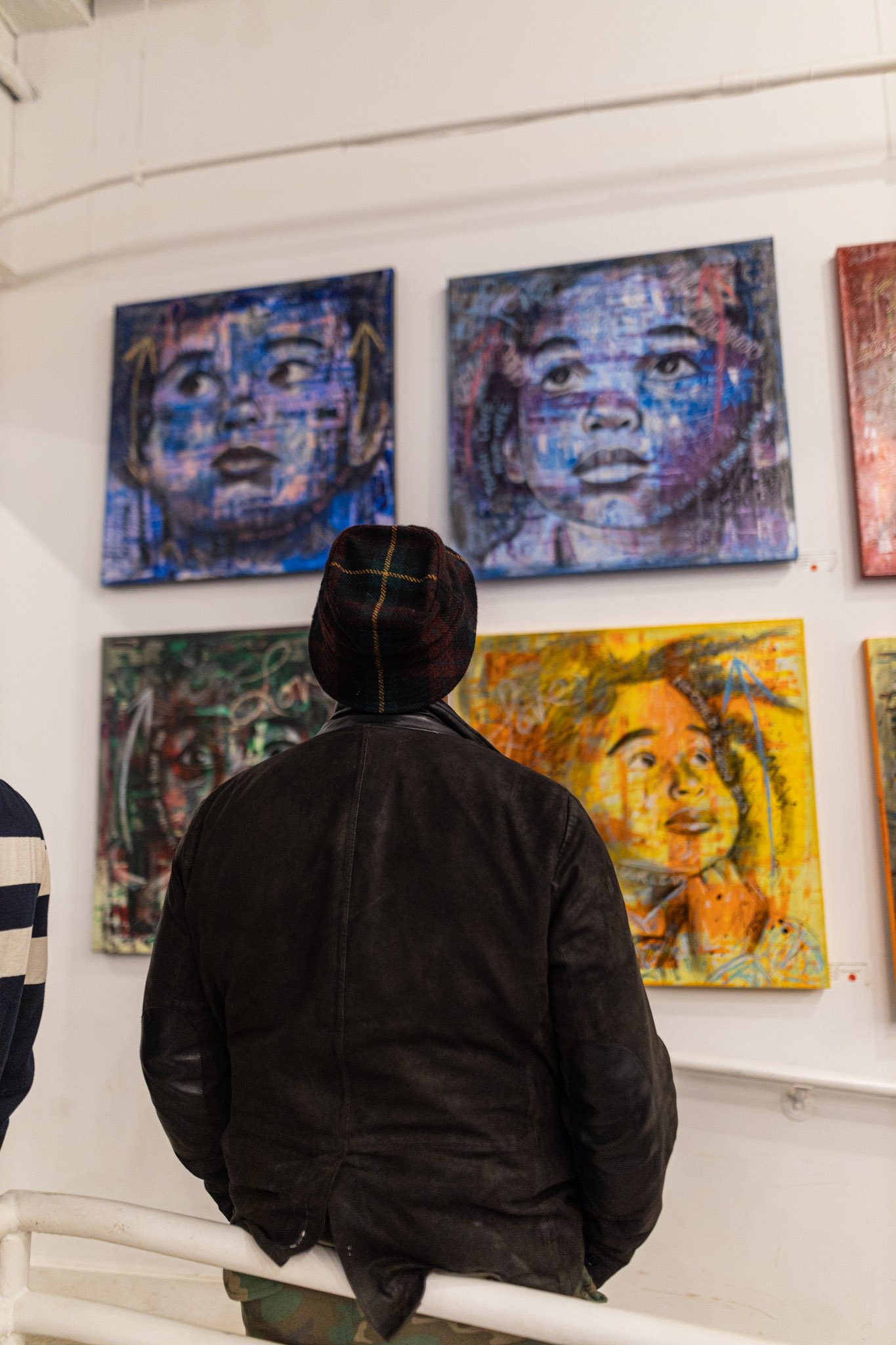 Person wearing a dark jacket and checkered hat standing and viewing colorful artwork of two children's faces at an art gallery