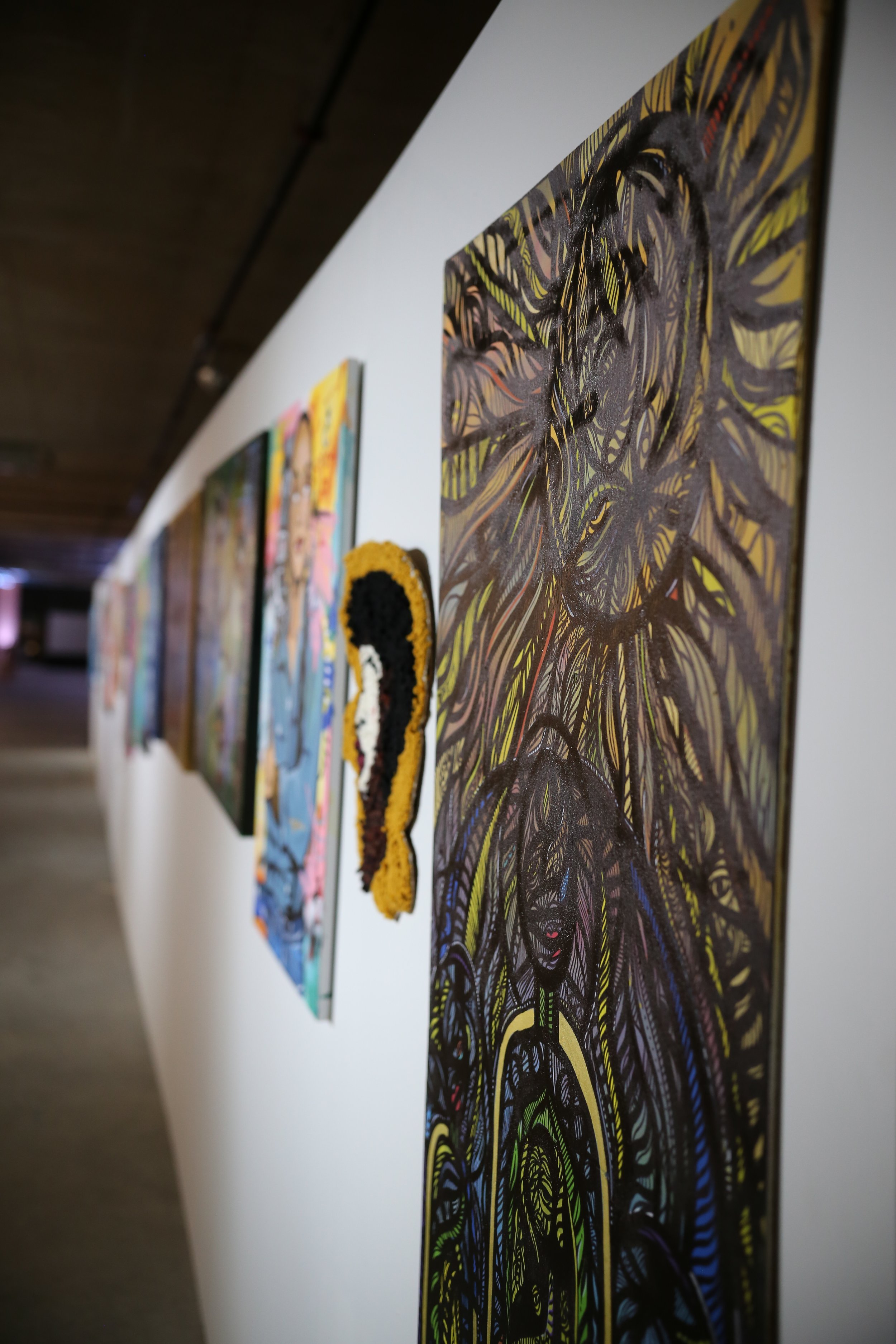 Art gallery wall with colorful abstract and illustrative paintings displayed in a row, viewed from an angle.