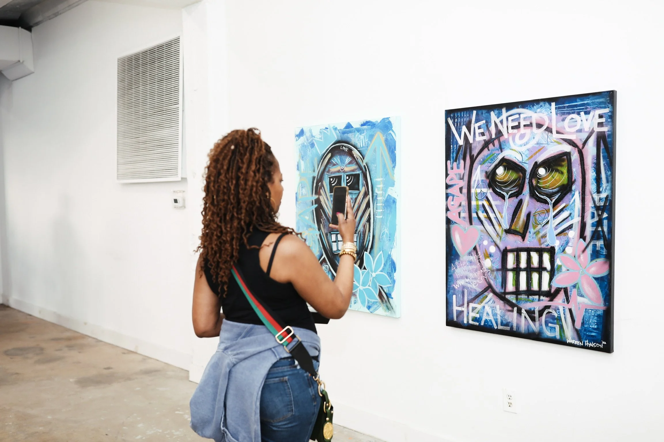 A woman with curly hair and a black tank top taking a photo of colorful graffiti art on a gallery wall, wearing a blue denim jacket tied around her waist and carrying a rainbow-striped shoulder bag.