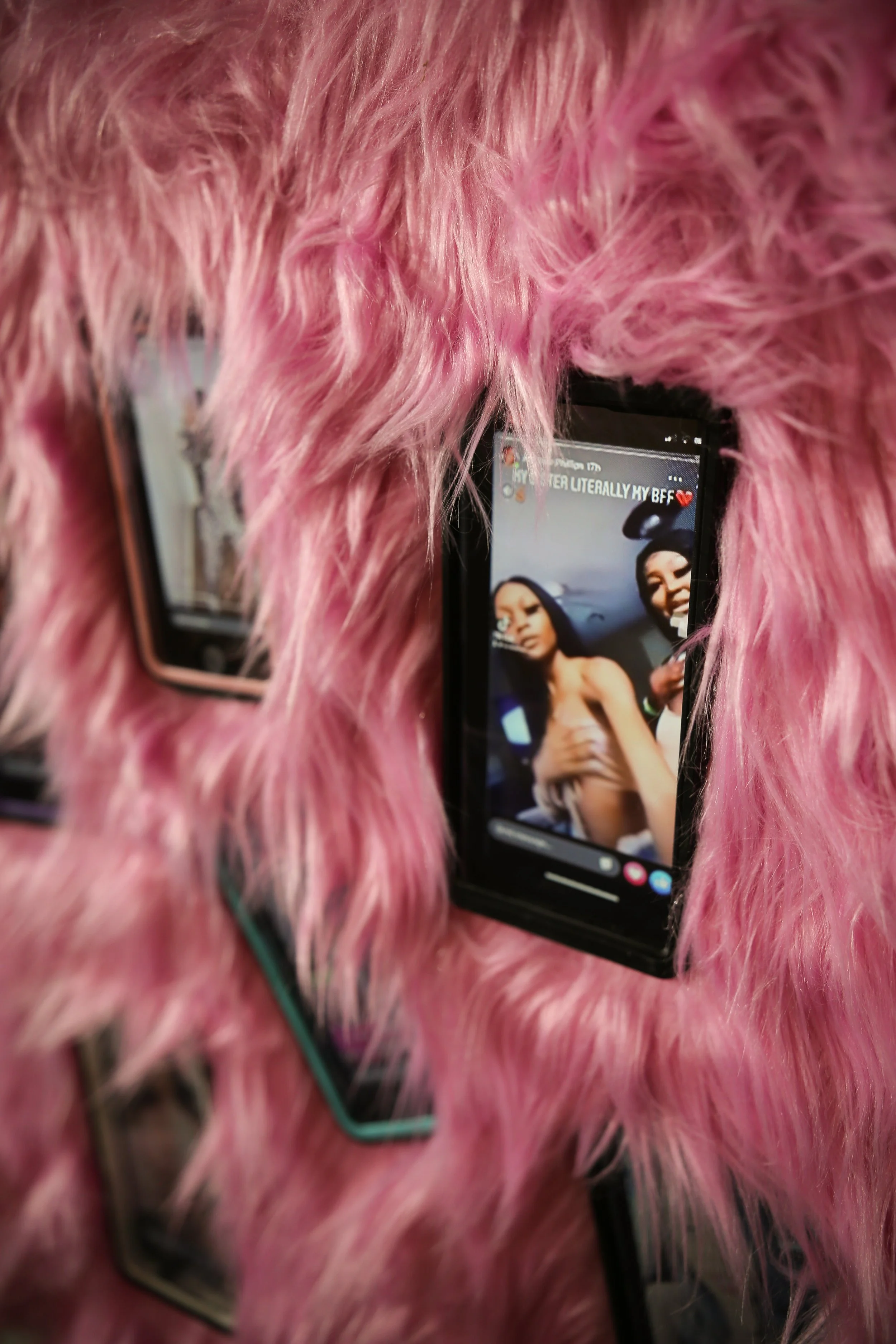Two smartphones are placed on a fluffy pink material, with screens displaying photos of women. One phone's screen shows a woman in a swimsuit, and the other shows a woman with dark hair. The pink material covers most of the phones.