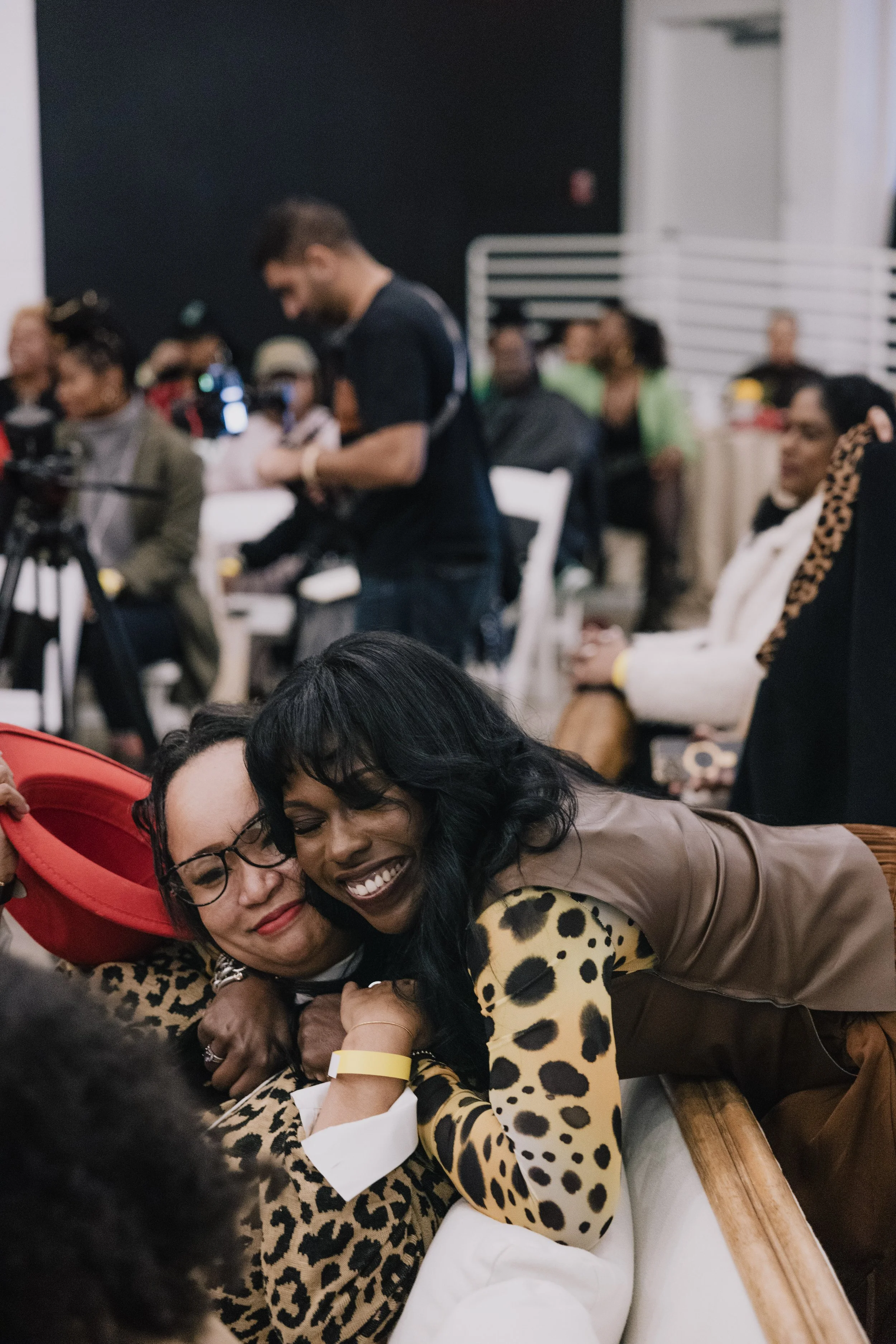 Two women hugging and smiling in the foreground, one wearing a red hat and the other in a leopard-print top, with a group of people blurred in the background, indoors at an event.