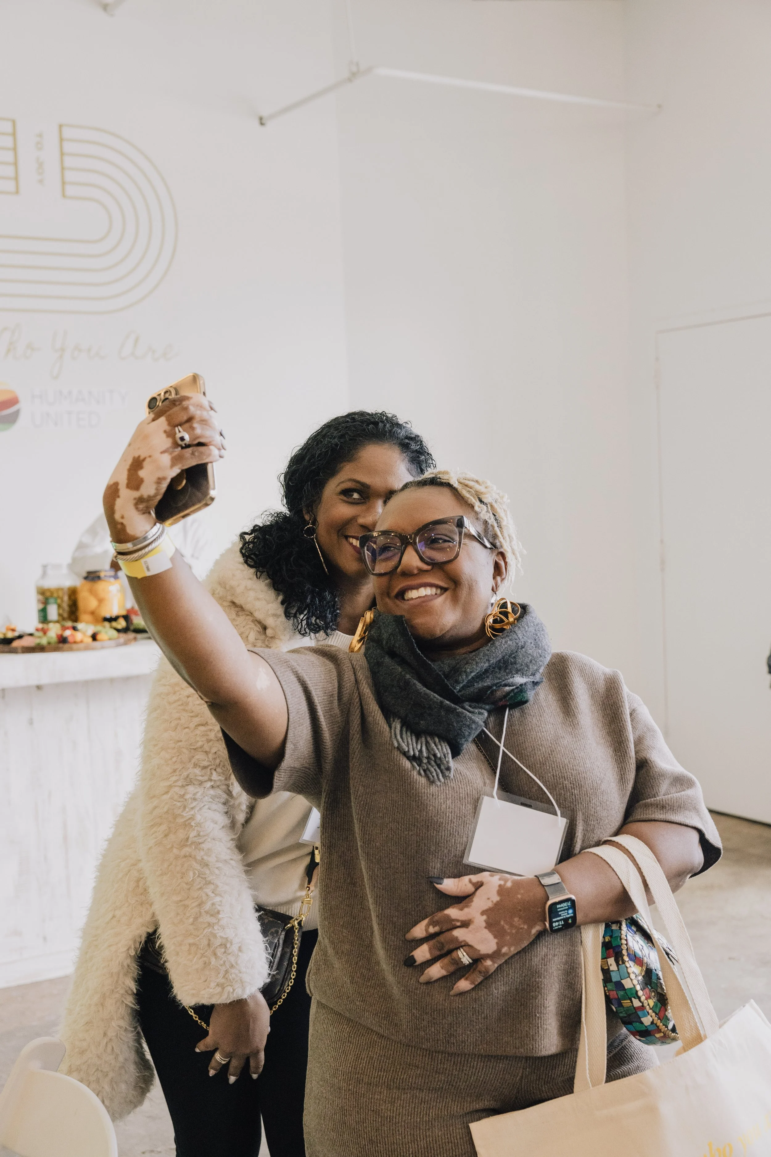 Two women smiling and taking a selfie at a social event with a white wall, some tables, and food in the background.