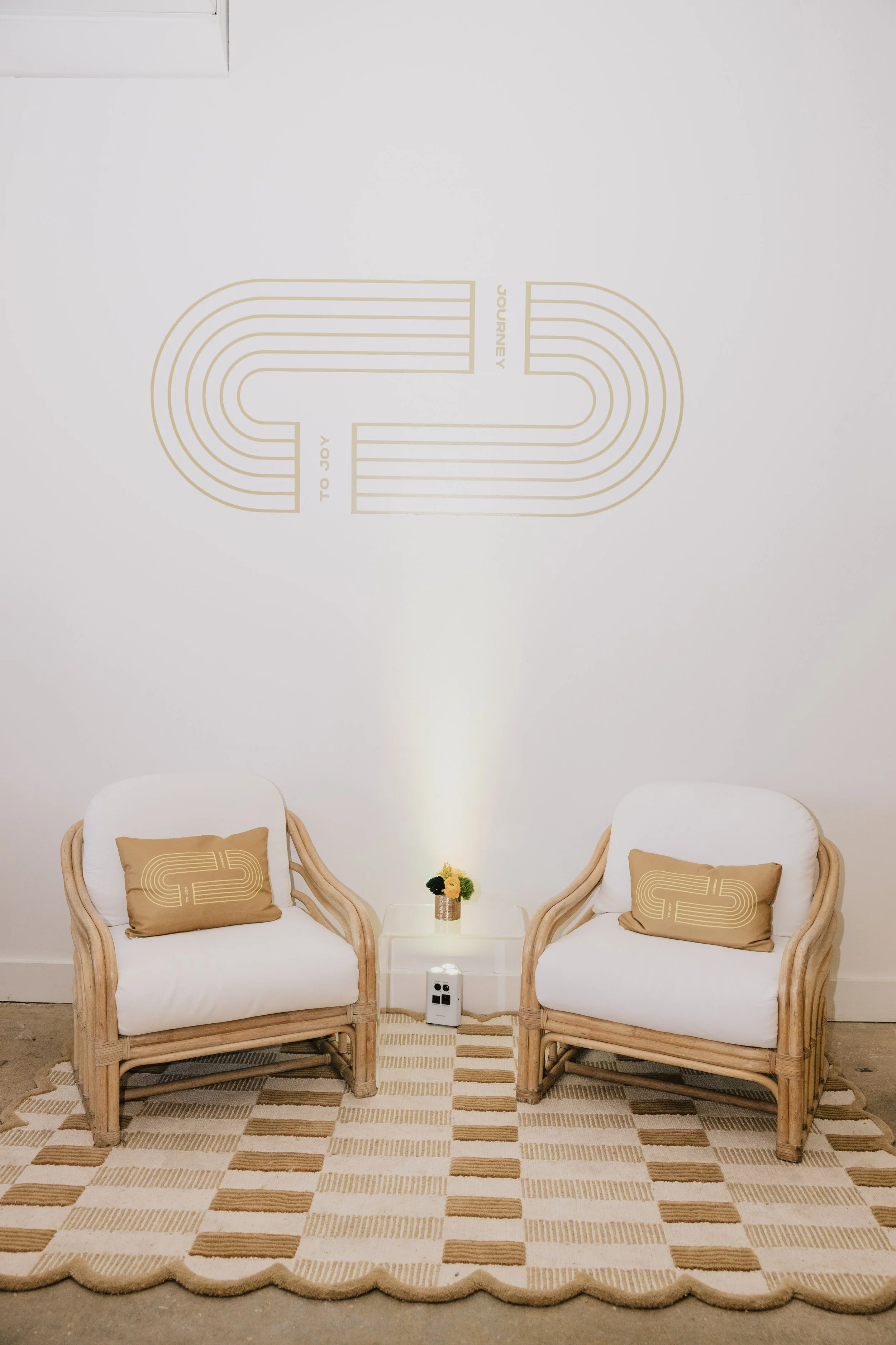 Two white chairs with beige cushions featuring gold outline designs, placed on a beige and white checkered rug with scalloped edges, a small white table with a potted plant, and a white wall with minimalist gold lines and words 'TO JOY' and 'JOURNEY' written on it.