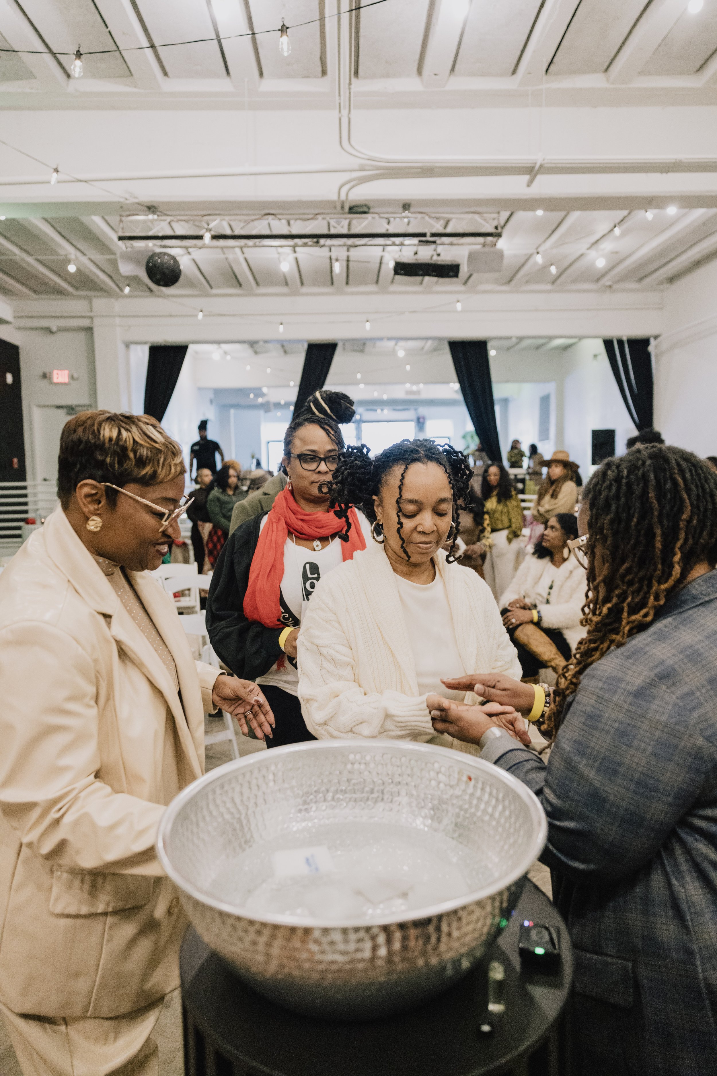 People gathered at an indoor event, with some engaging in a ceremonial activity, such as a prayer or blessing, around a large bowl on a table.