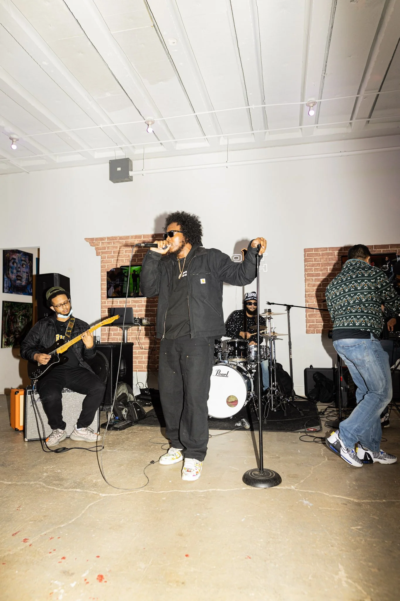 A musical band performing live on a stage in an indoor venue with brick wall decorations and artwork. The band consists of a singer, a guitarist, a drummer, and a keyboard player.
