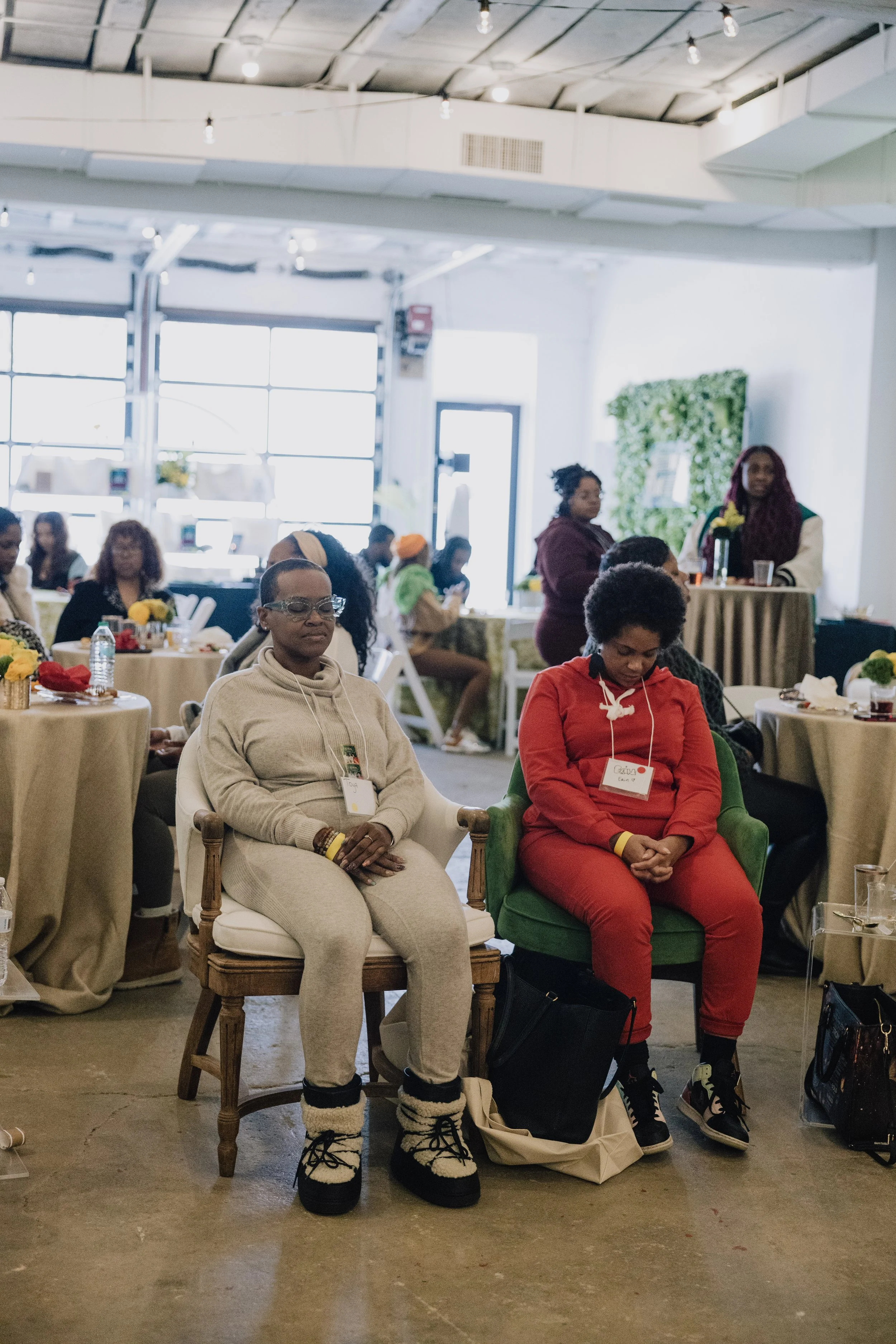 People attending an indoor event, seated at round tables with floral centerpieces, with some individuals wearing name tags. Two women are sitting in the foreground, with their eyes closed, one dressed in beige and the other in red. The background shows more attendees, some standing and some seated, in a well-lit space with large windows and a green plant wall.