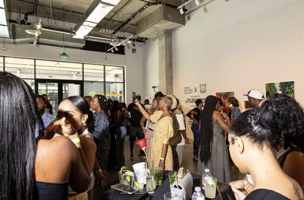 Crowd of people at an art gallery exhibition, viewing artworks on the walls, sitting at tables, and taking photos.