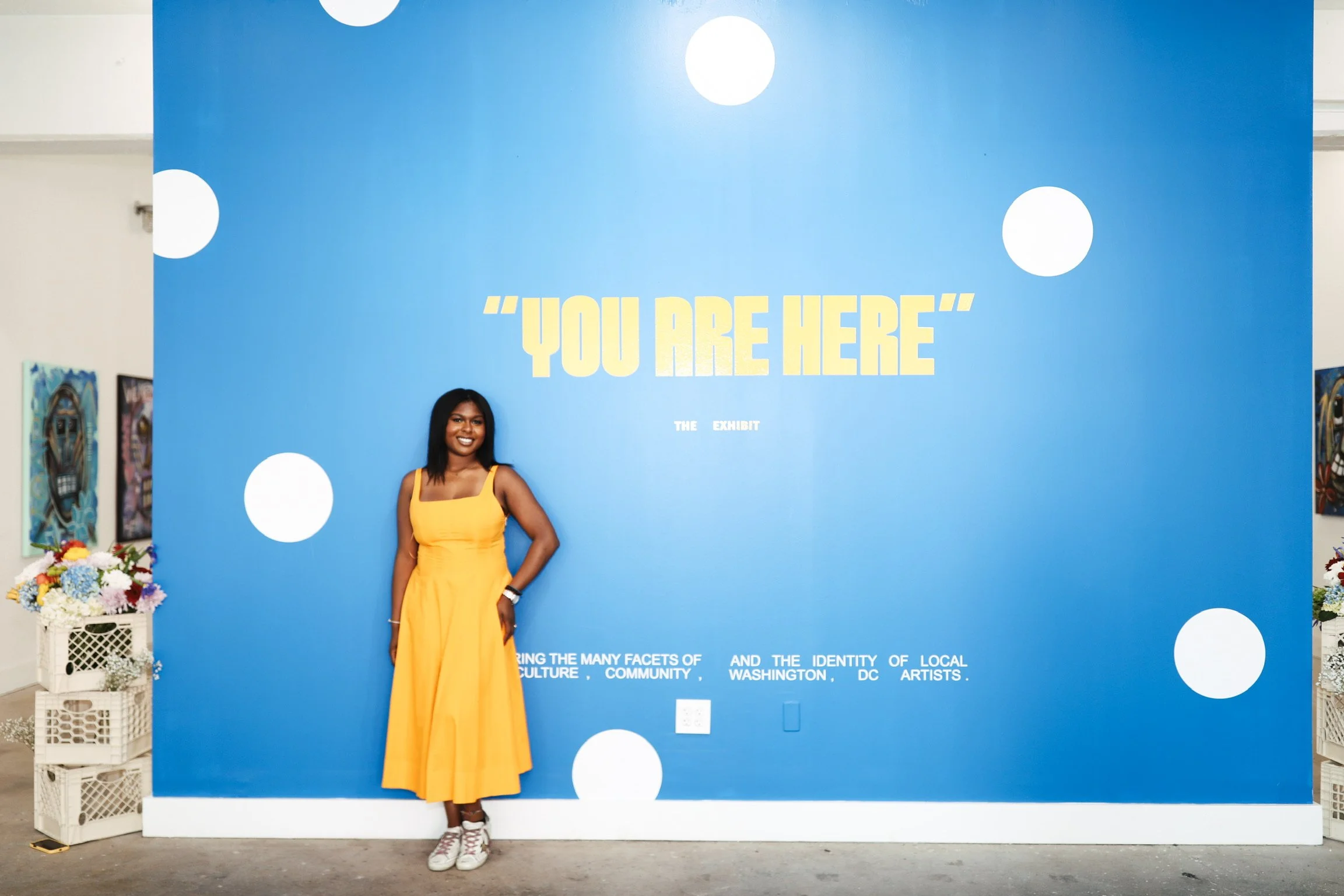 Woman in yellow dress standing in front of blue exhibition wall with the quote "YOU ARE HERE" and white polka dots.