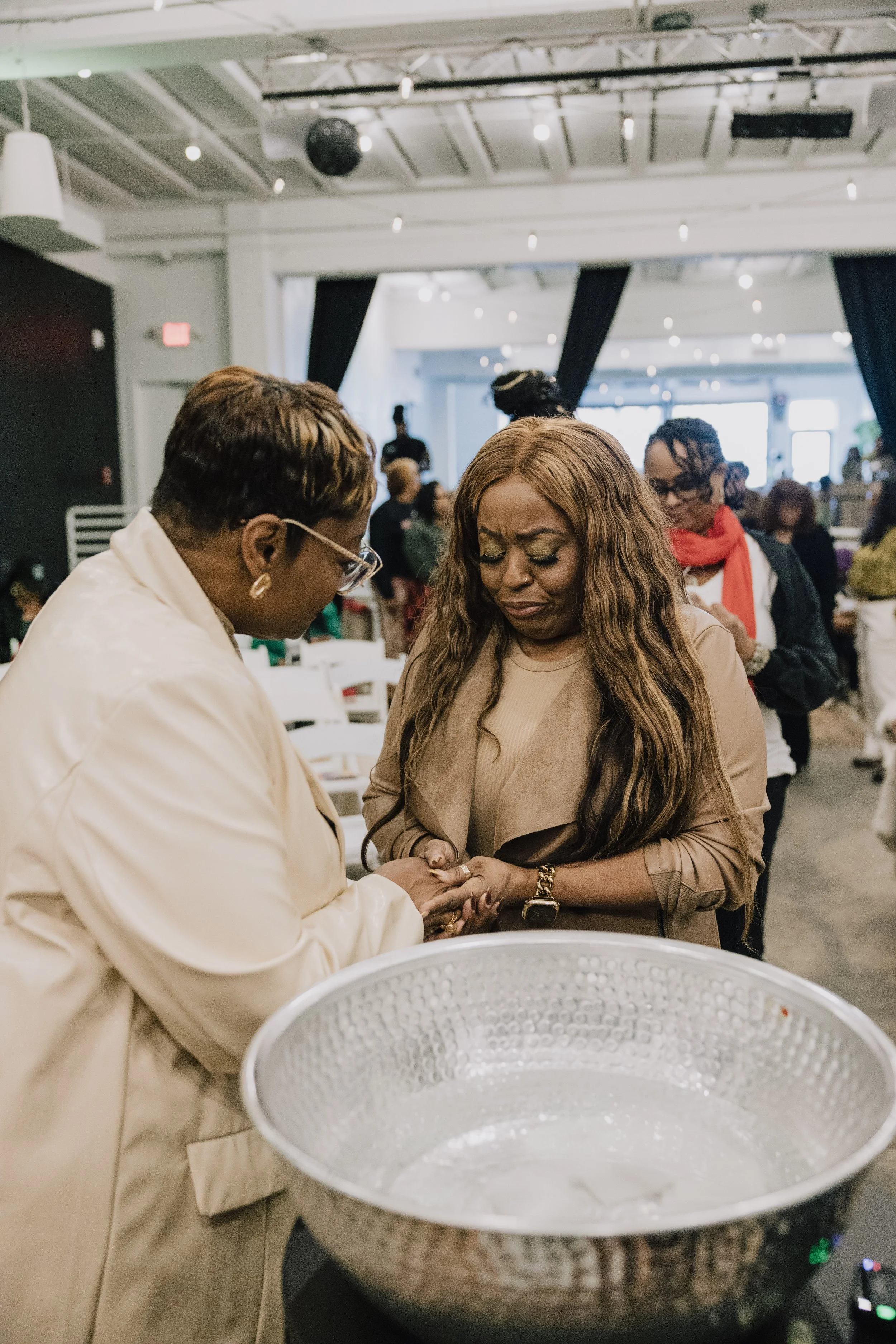 Two women holding hands, standing in a bright, spacious indoor setting with other people in the background. One is wearing glasses and earrings, and the other has long wavy hair and a watch. There is a large metallic bowl in the foreground.