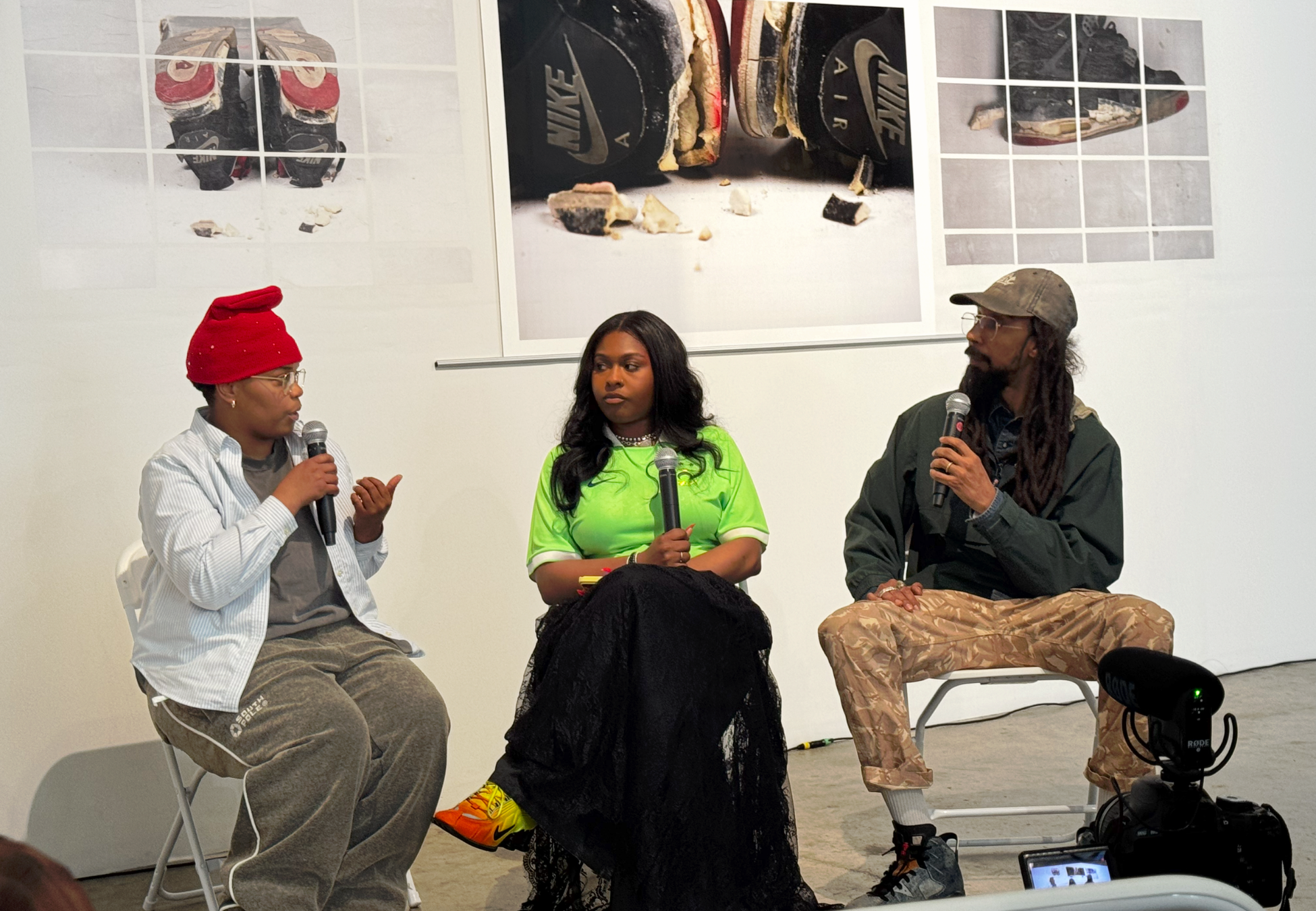 Three people sitting on chairs, participating in a panel discussion or interview, with microphones in hand, against a white wall with photographs of sneakers.