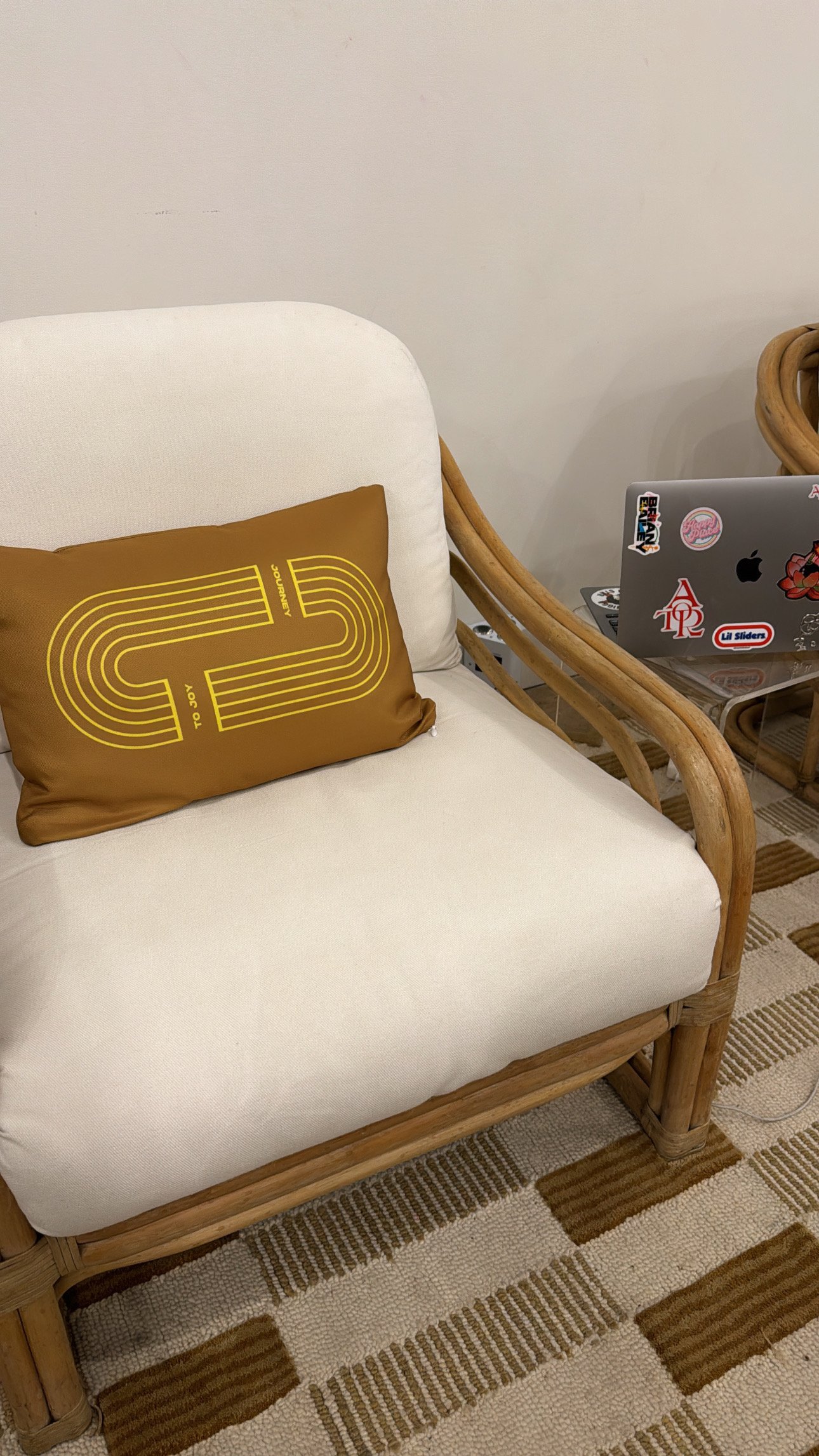 A wicker sofa with a white cushion and a brown pillow with yellow text. Next to it is a glass table with a laptop covered in stickers. The setting appears to be in a cozy indoor space with a beige wall and a patterned rug.